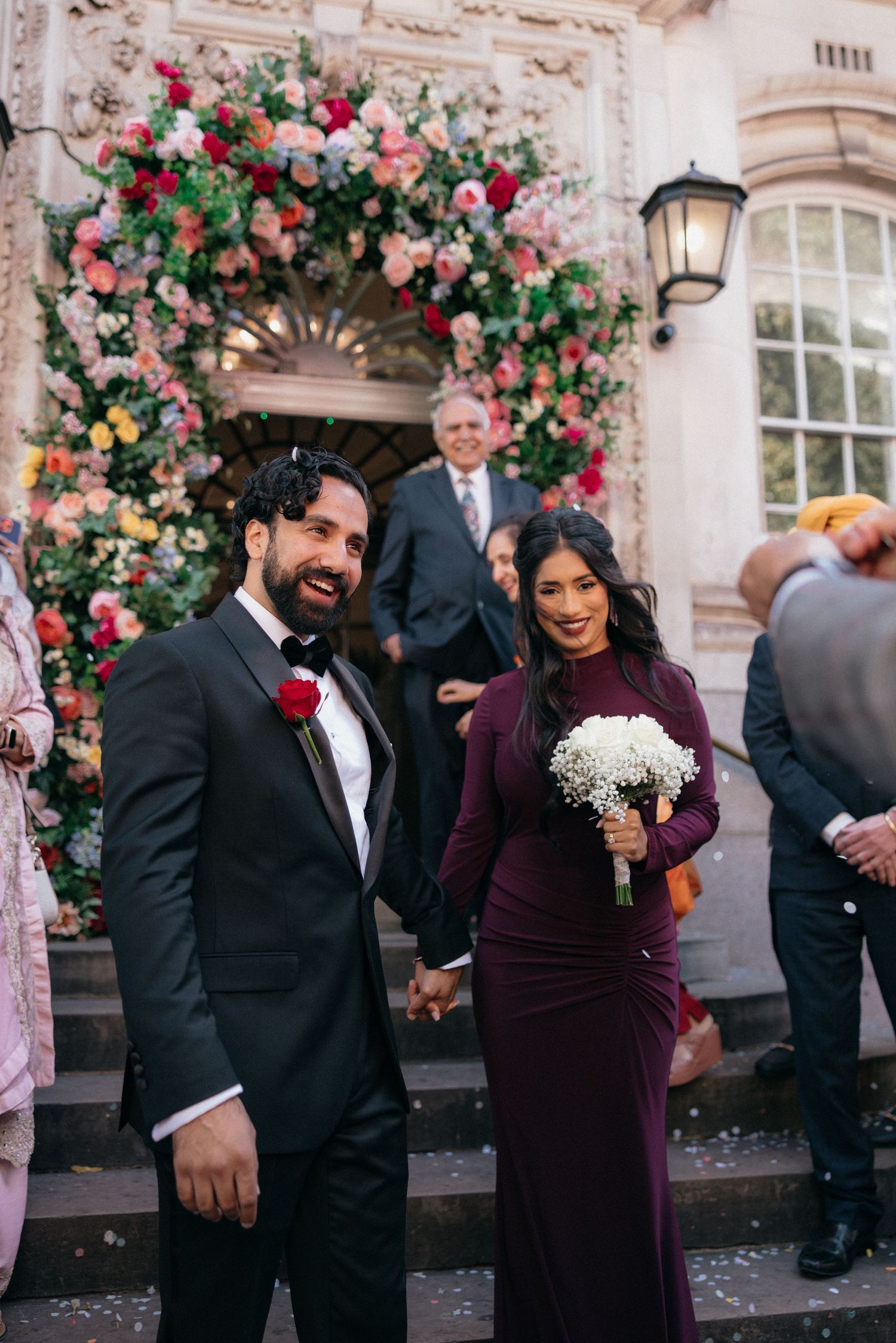 Wedding party poses under beautiful pink and red rose floral arch at elegant venue entrance.