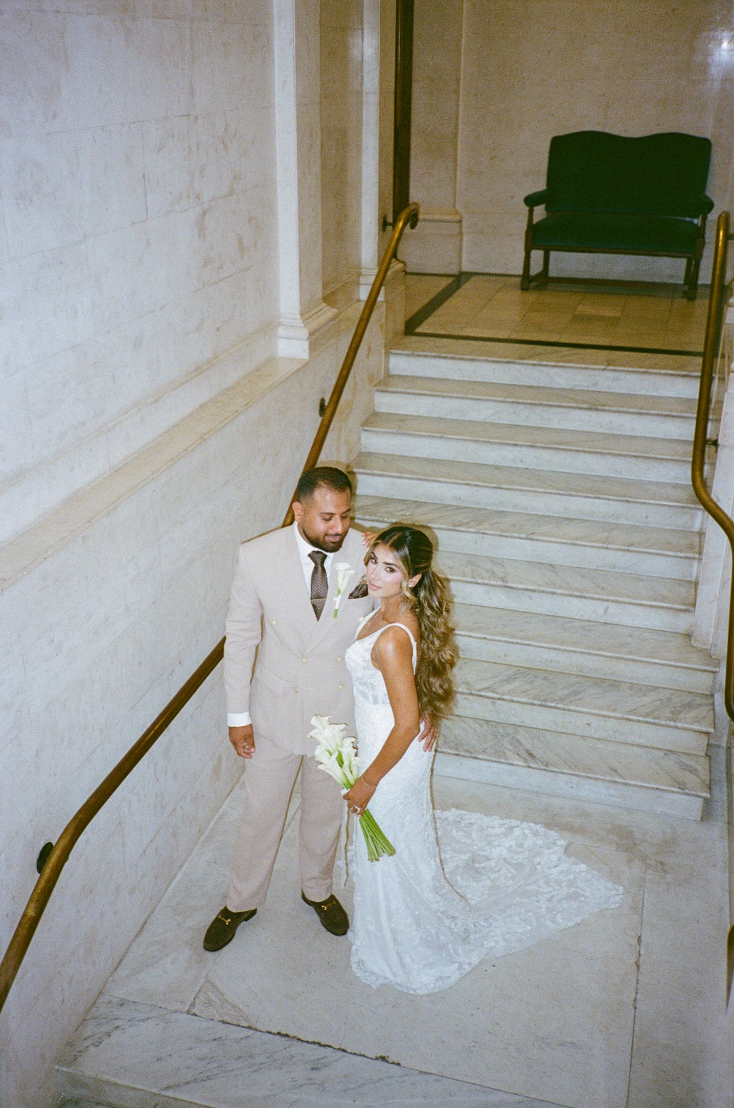 Bride and groom pose on marble staircase, she holds white bouquet in lace gown, he wears beige suit.