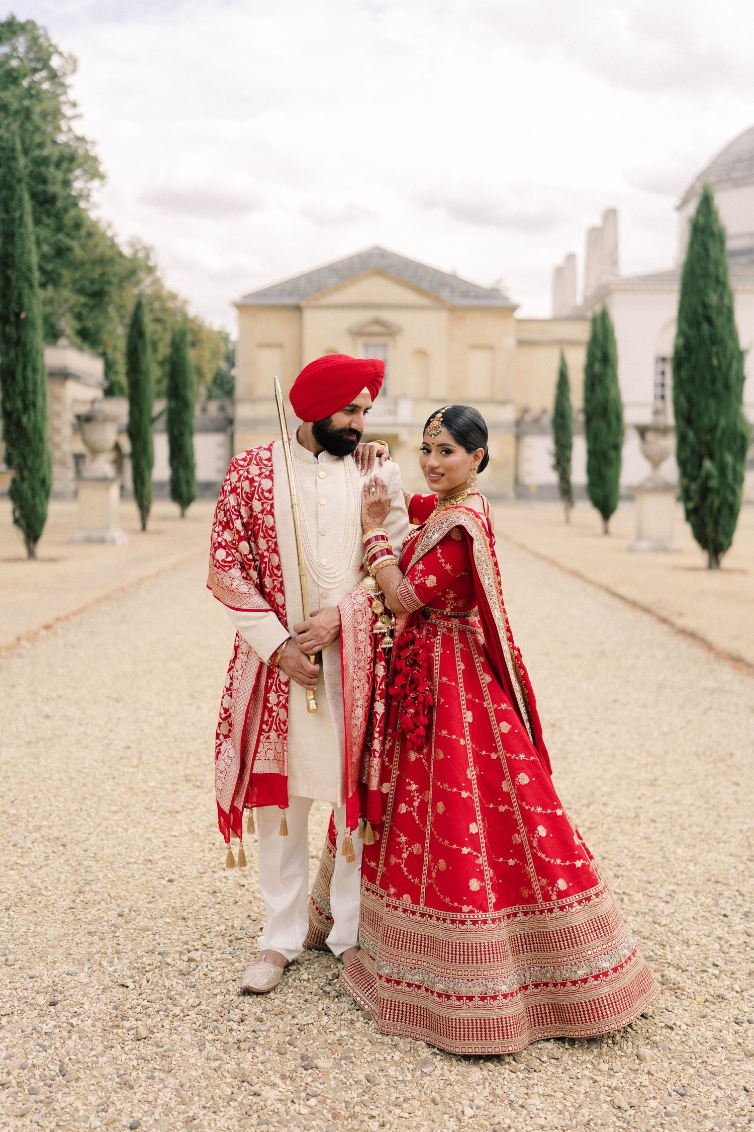 Sikh bride and groom in traditional red and gold wedding attire pose on a gravel path at an elegant estate.