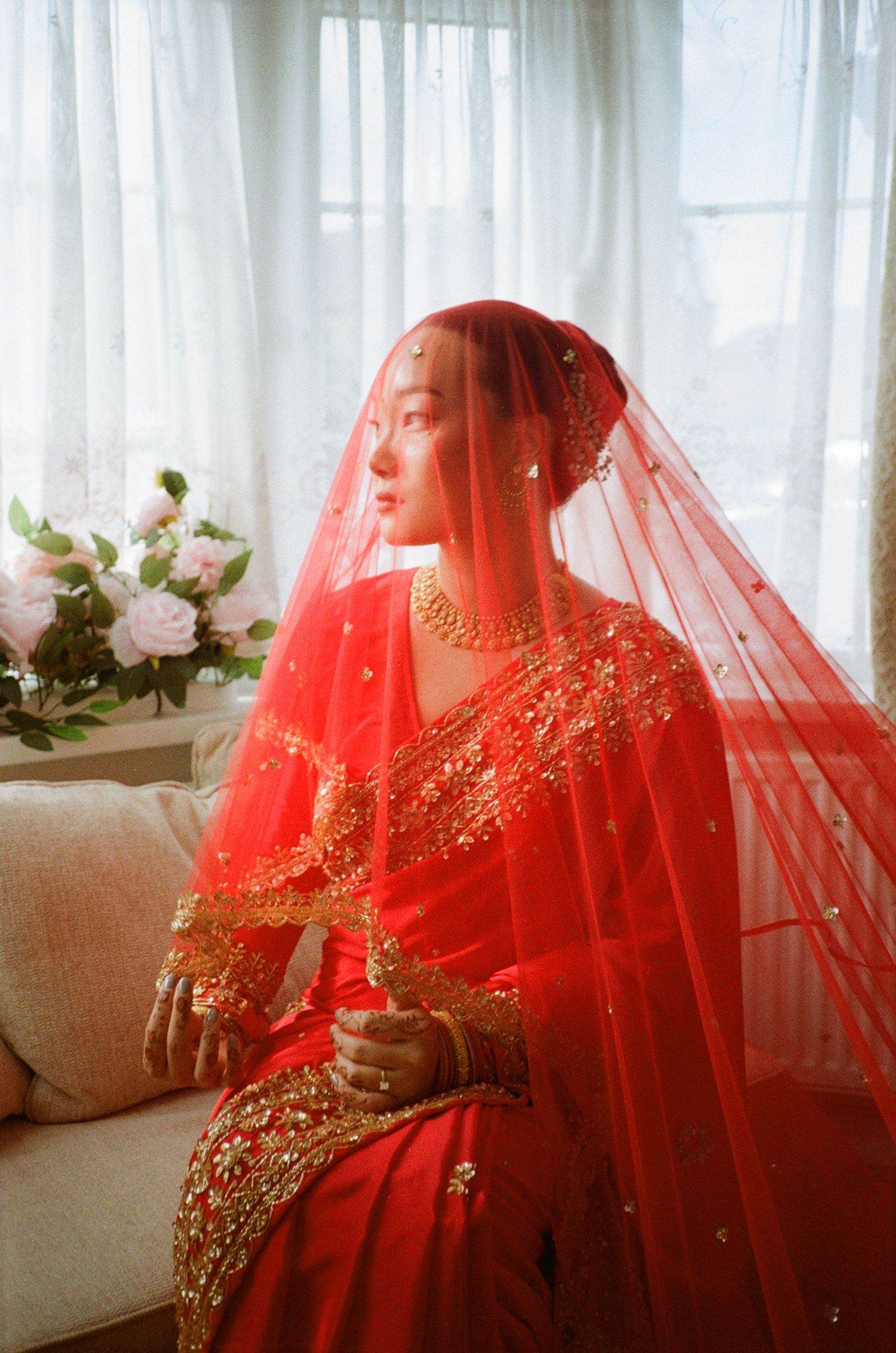A bride in a red saree and gold jewelry sits gracefully, wearing a red veil, with flowers in the background.