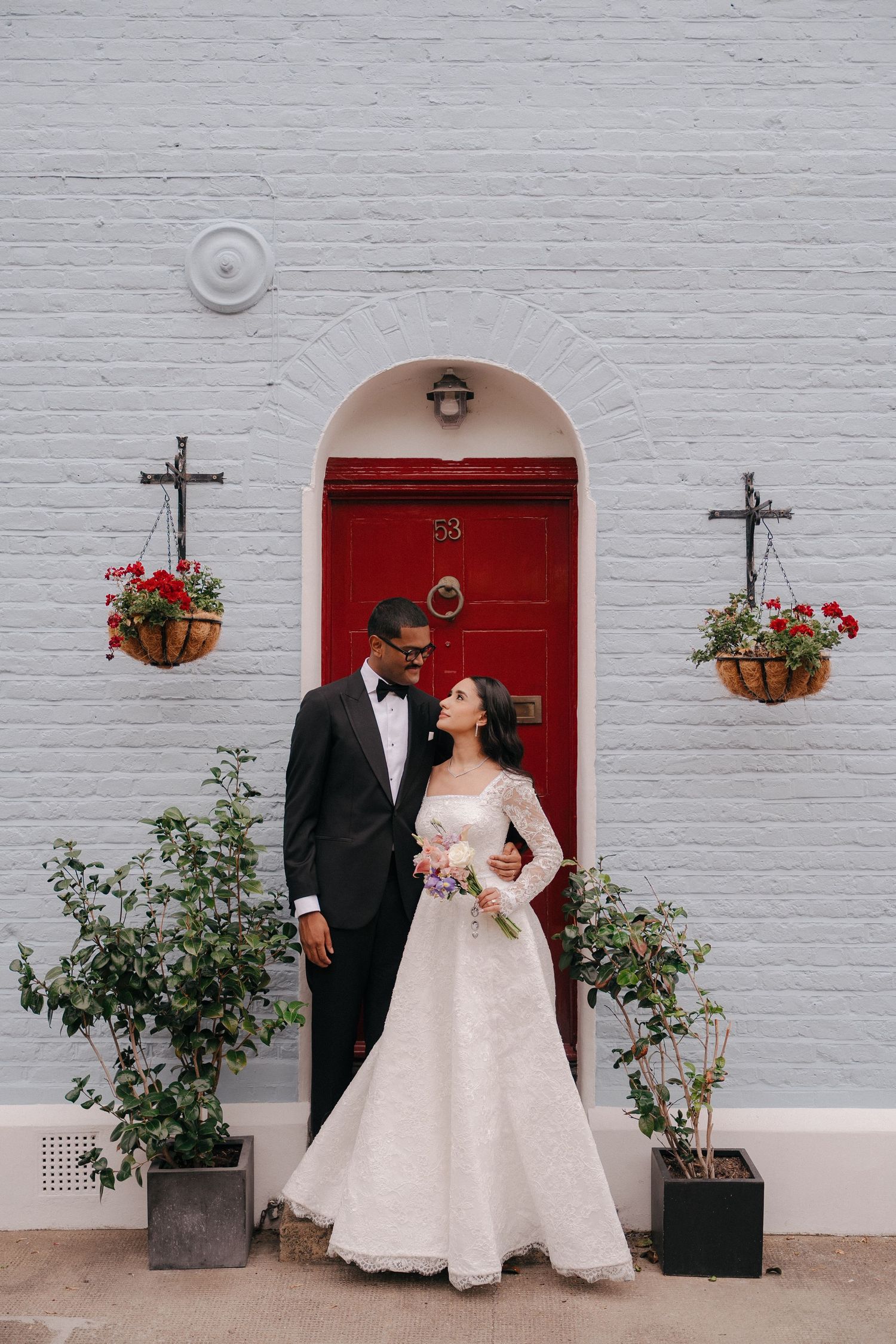 Wedding couple poses against white brick wall with red door and climbing roses.