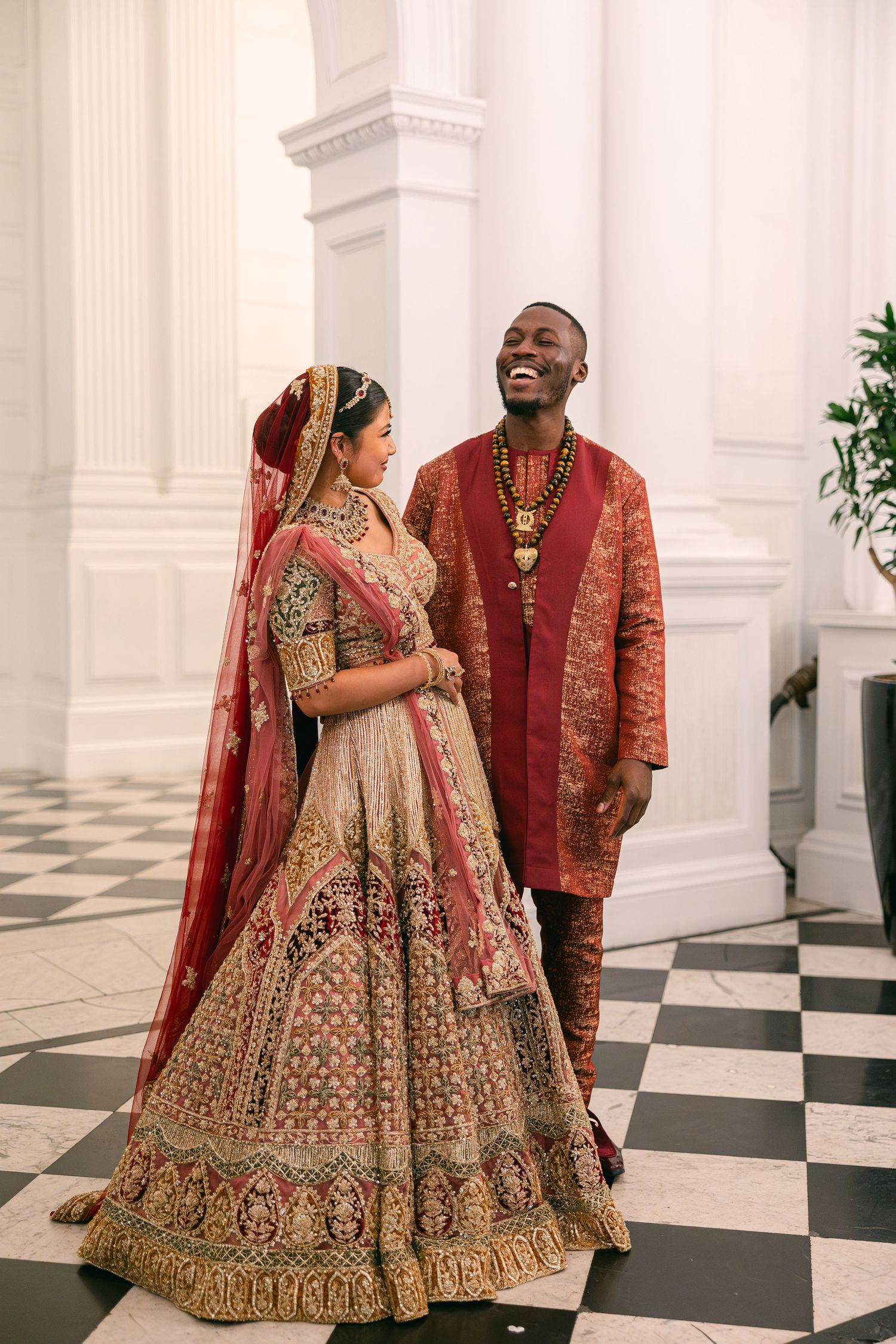A couple shares joyful moments in their ornate red and gold South Asian wedding attire against white columns.