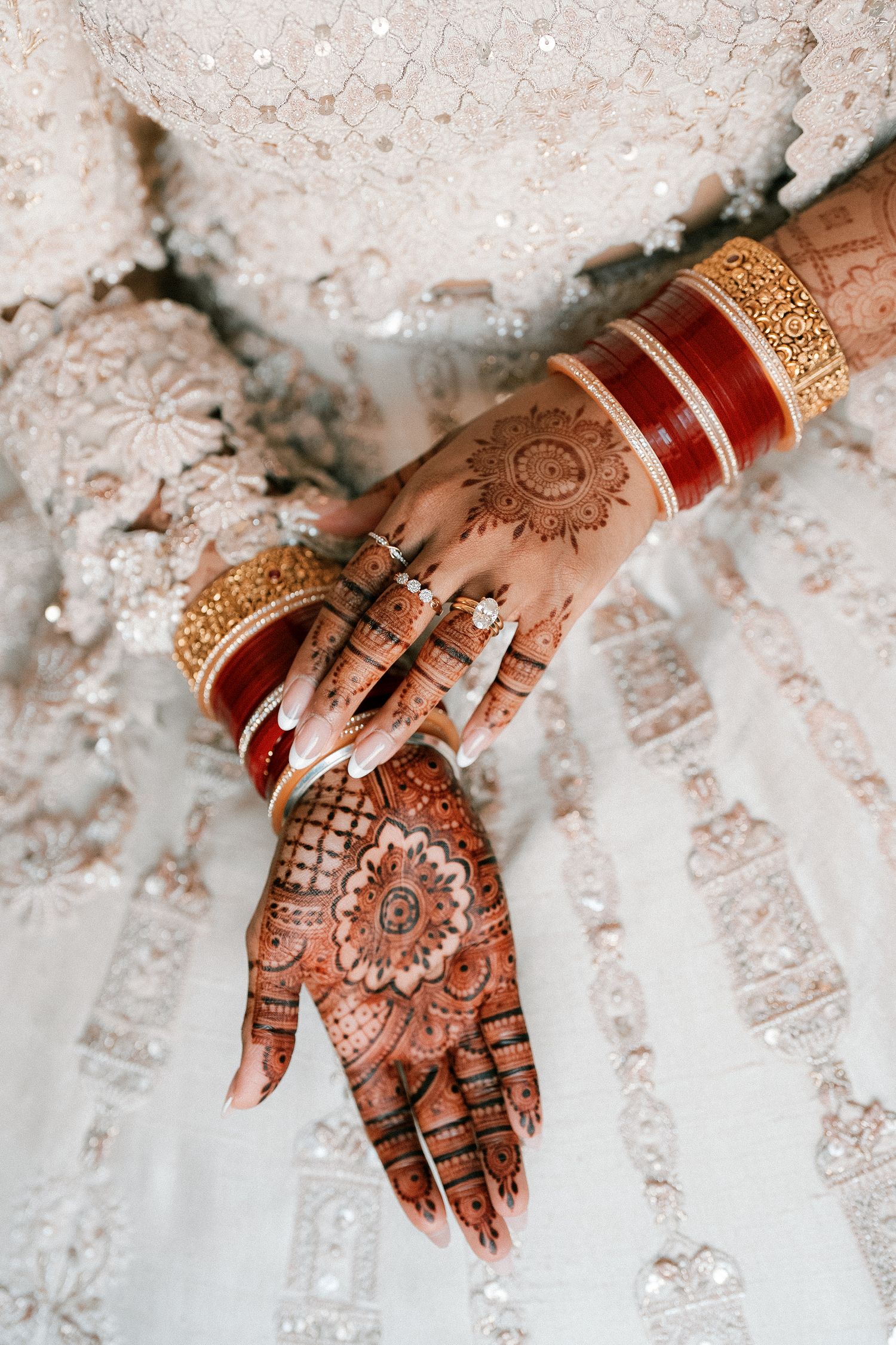 Bride's hands adorned with intricate henna mehndi designs, gold and red bangles, and a diamond ring on white lehenga.