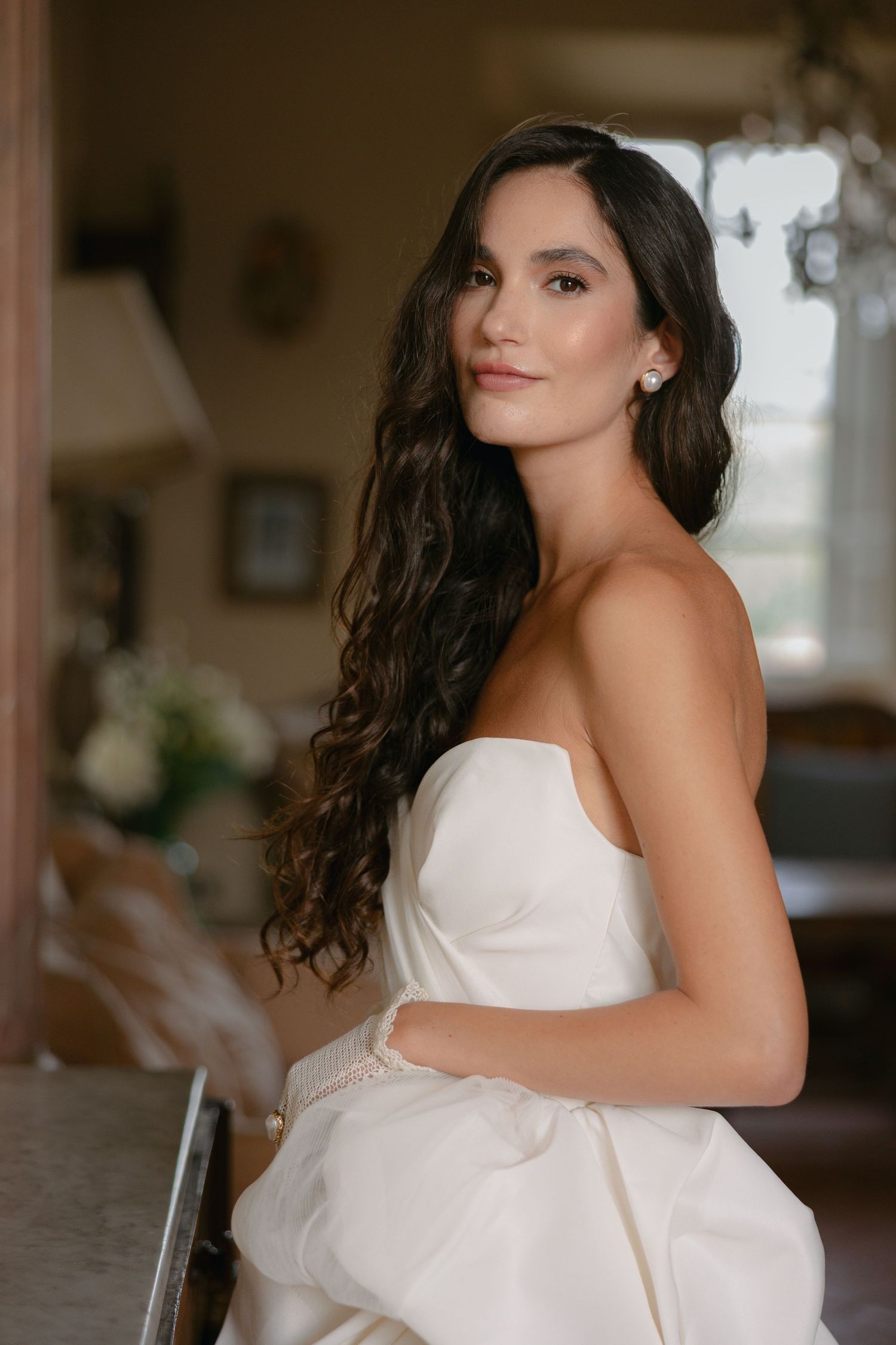 Bride in strapless white gown with long wavy brunette hair and pearl earrings posing elegantly indoors.