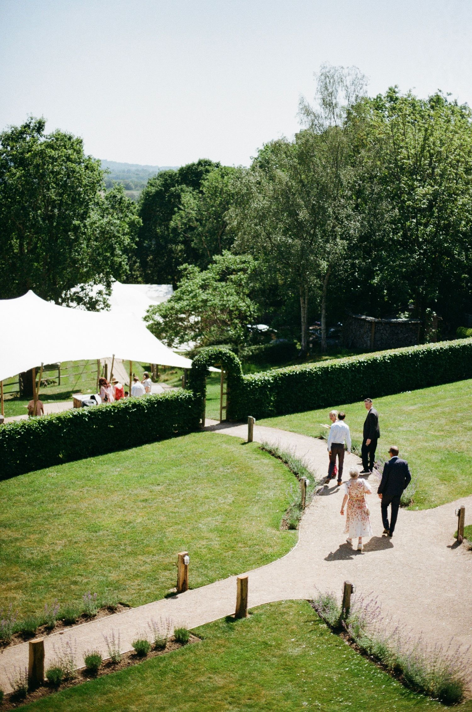 Guests walking along a curved garden path toward a white marquee tent at an outdoor summer wedding venue.