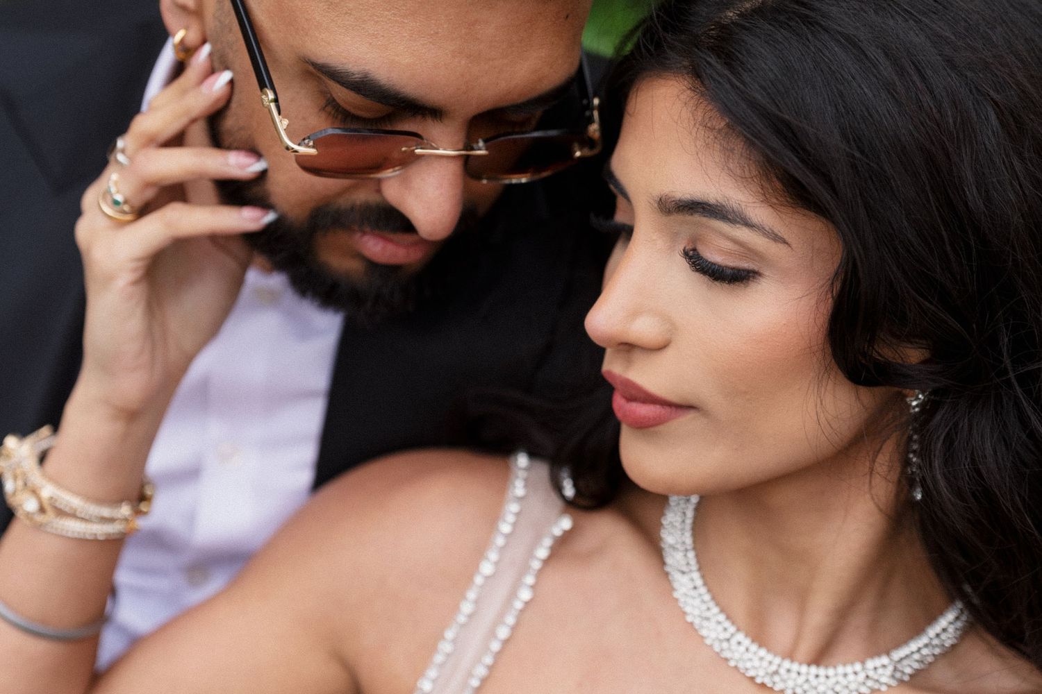 Romantic close-up of a South Asian couple in elegant jewelry, man in sunglasses leaning toward woman in pearl necklace.