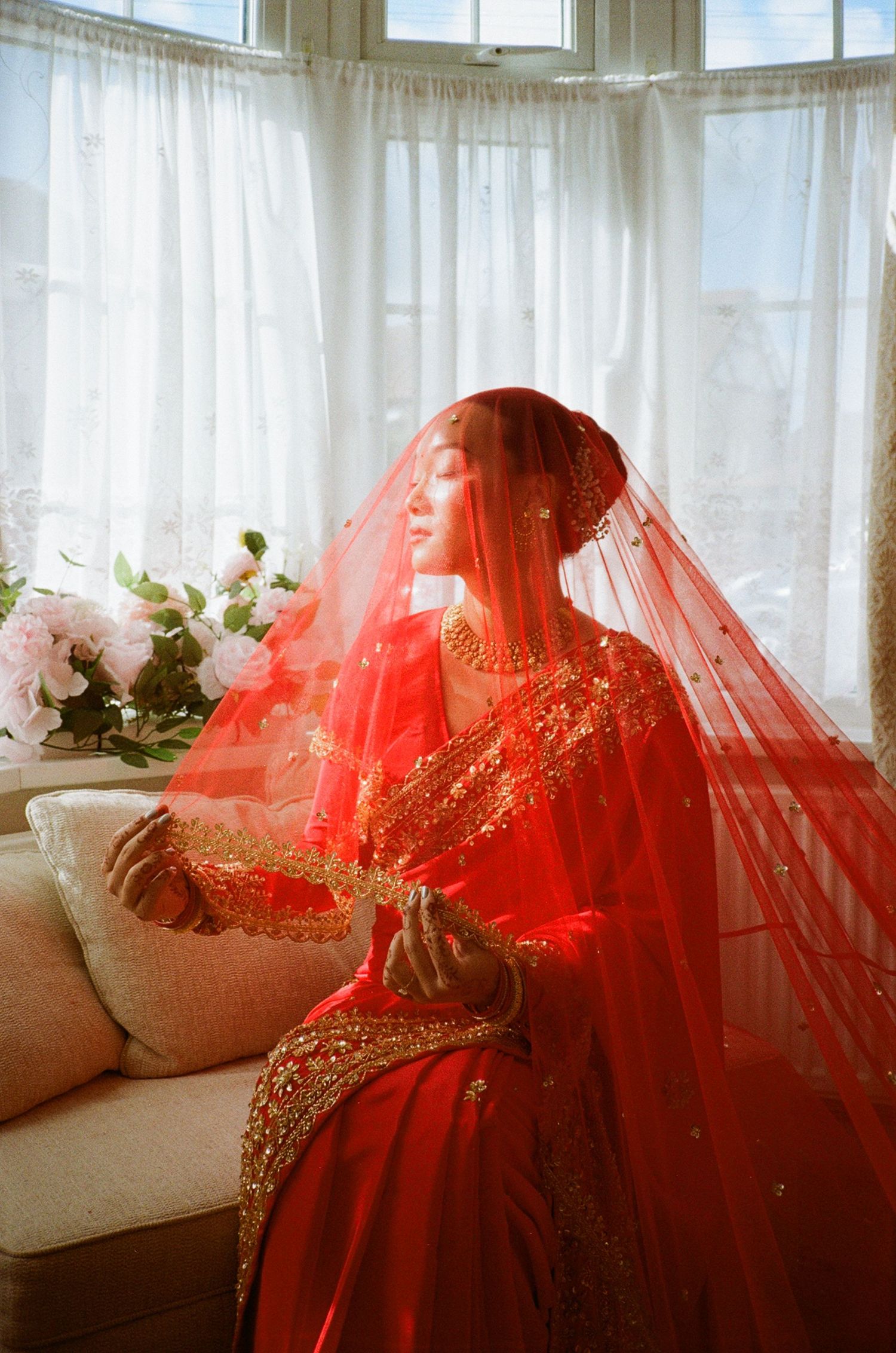 A bride in a red embroidered lehenga and gold jewelry sits gracefully by a sunlit window wearing a flowing red veil.