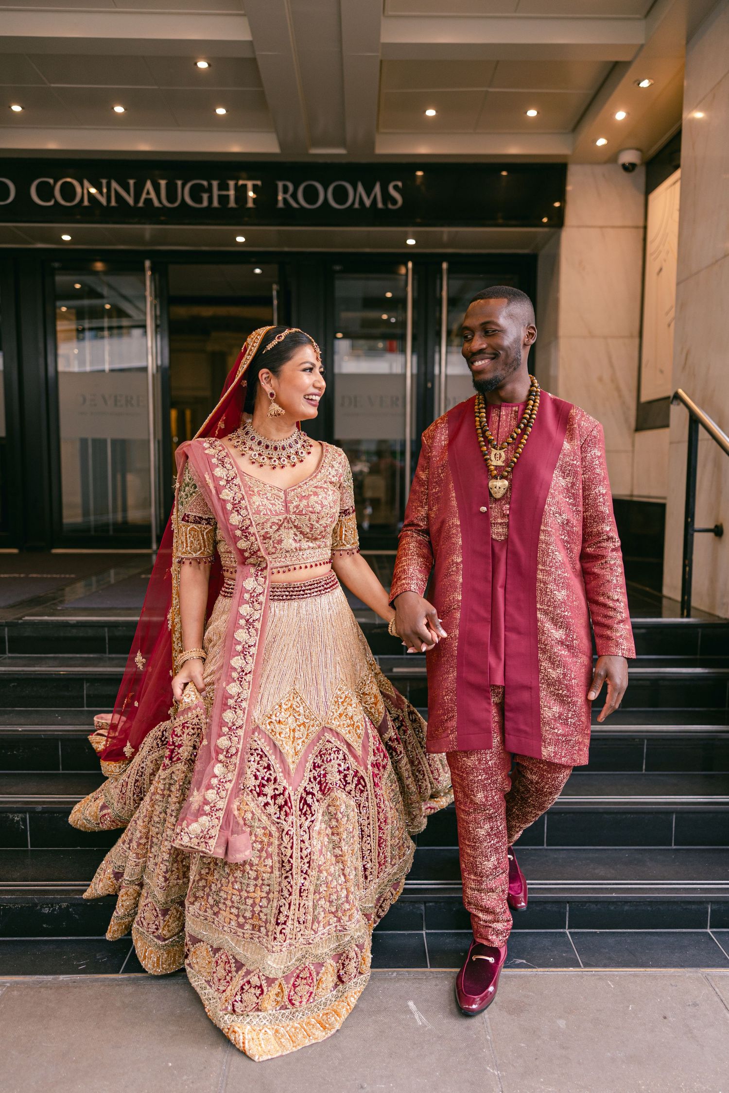Couple in coordinated pink and gold traditional wedding attire outside Connaught Rooms.