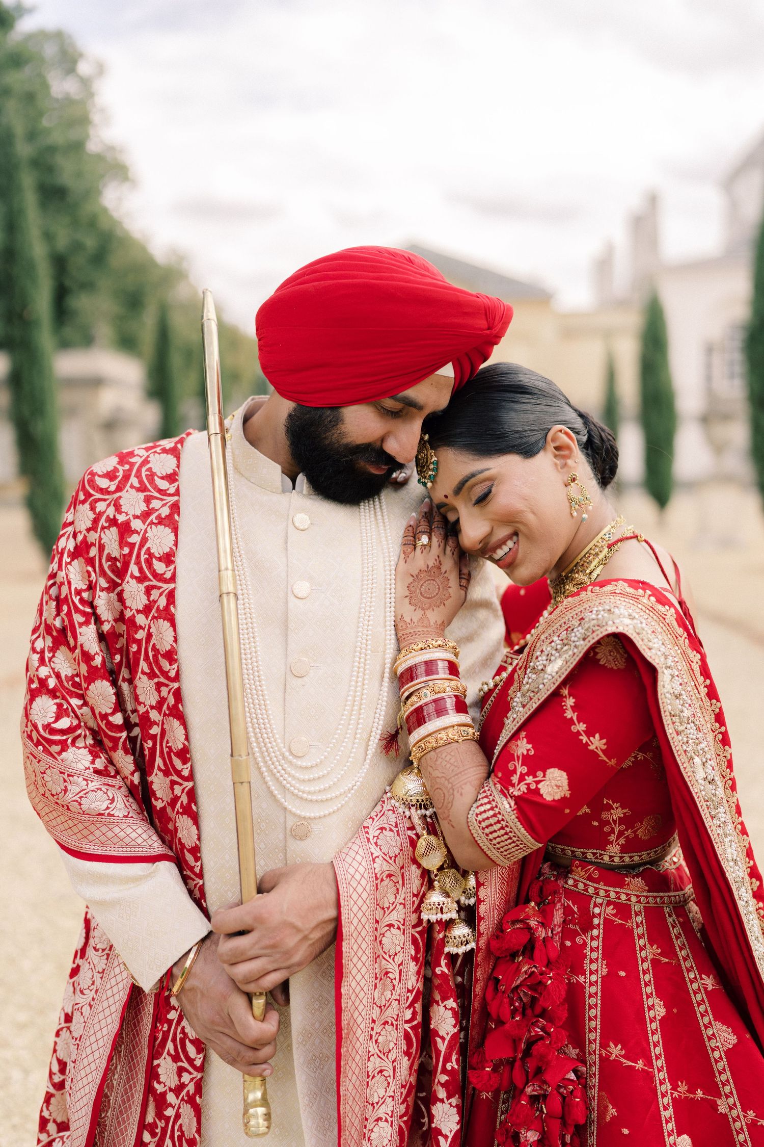 Sikh bride and groom in traditional red wedding attire share a tender moment outdoors.