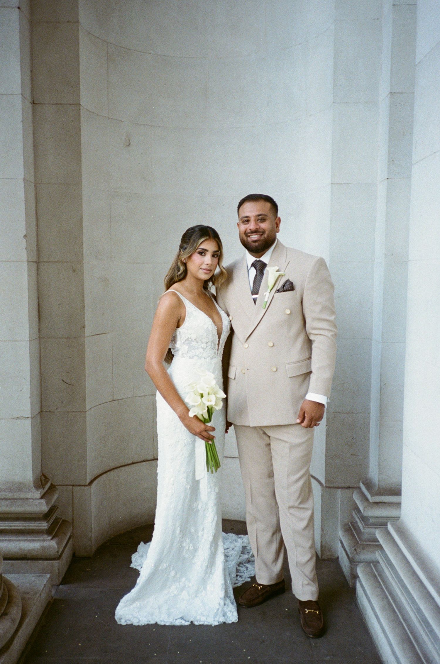Bride in white lace gown holding calla lilies bouquet stands with groom in beige suit beside stone columns.