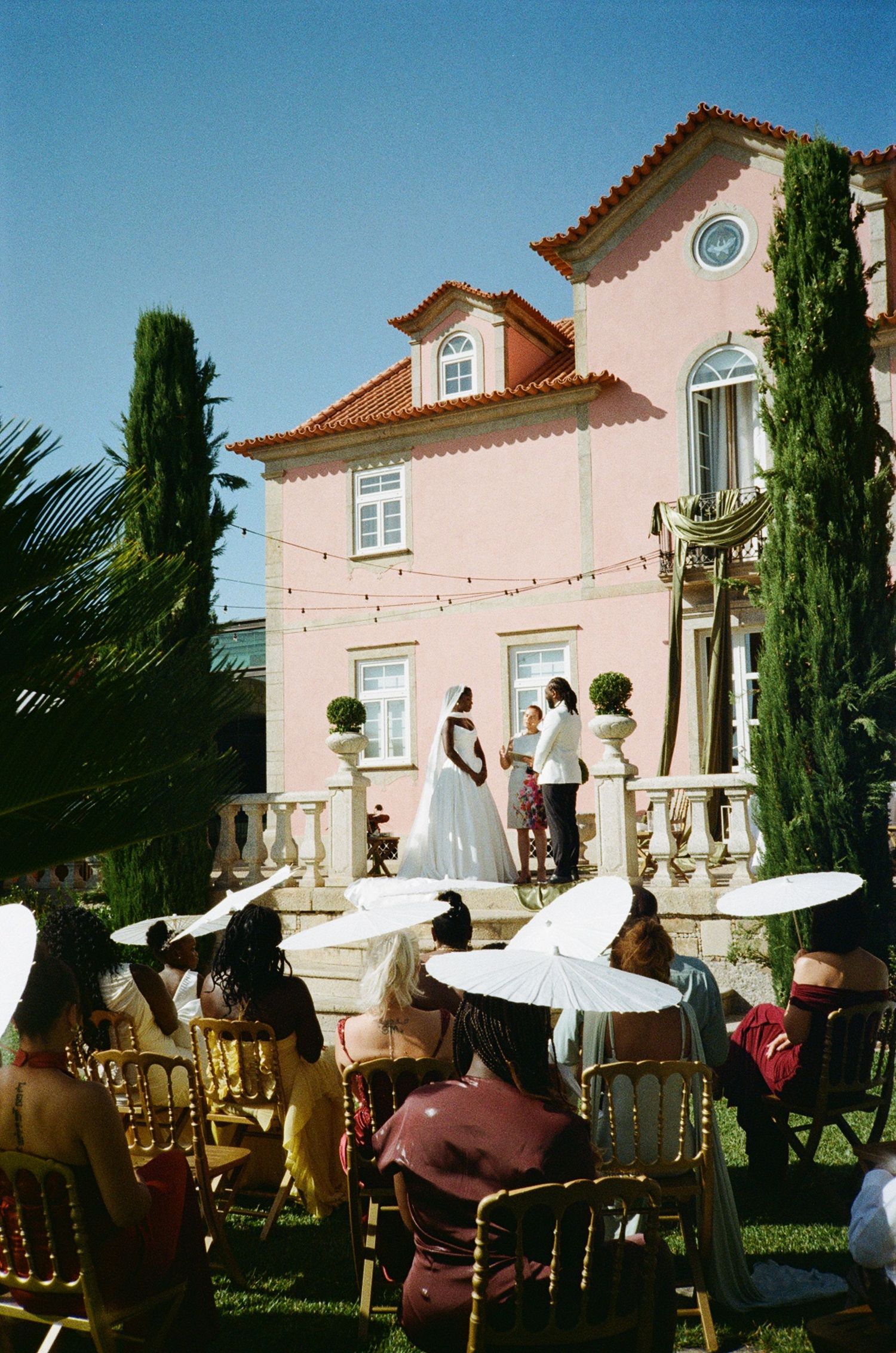 Outdoor wedding ceremony at a pink villa with guests seated in gold chairs holding white parasols under a clear blue sky.