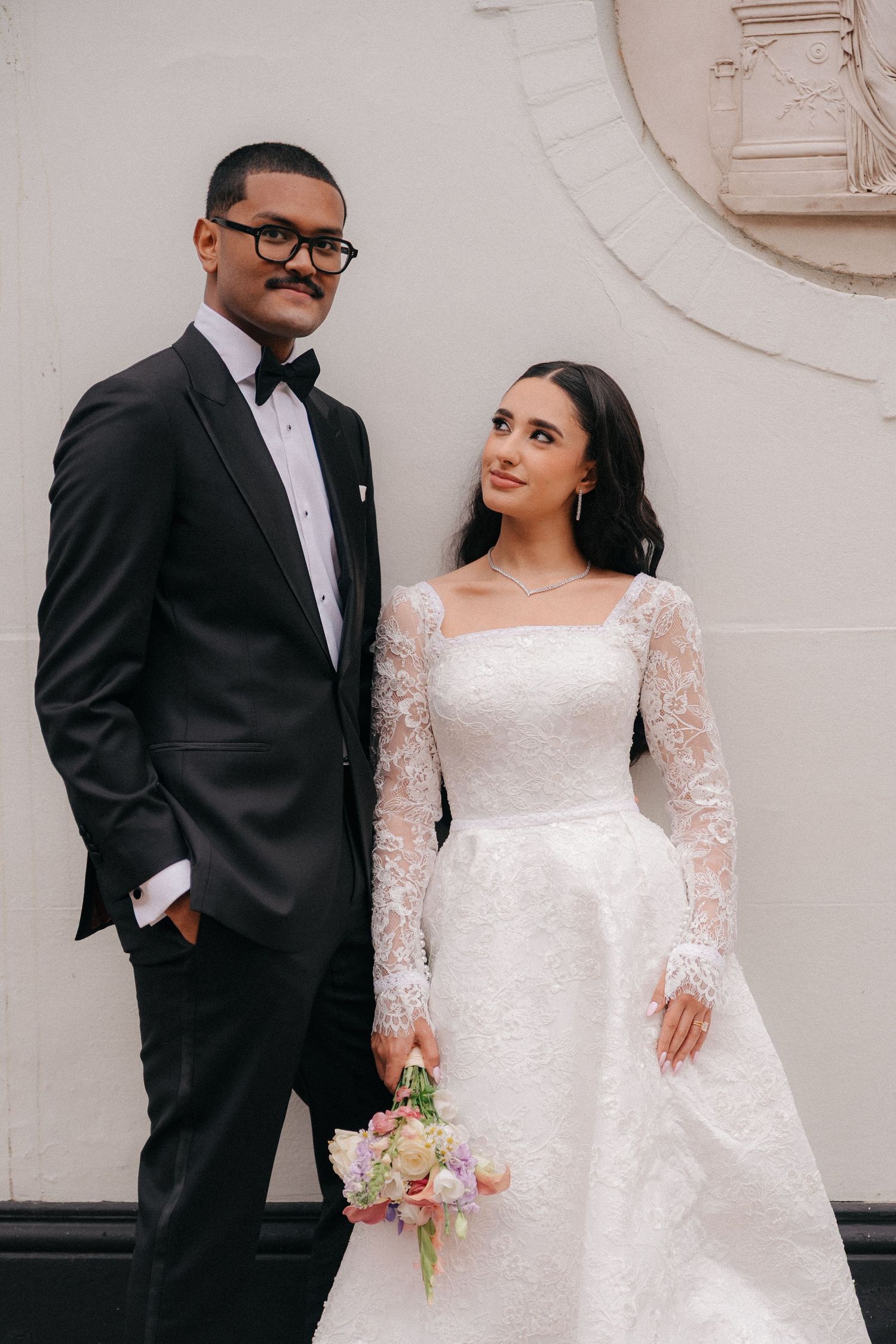 A wedding couple poses against a white wall, with the bride wearing a lace-sleeved gown and groom in classic tuxedo.