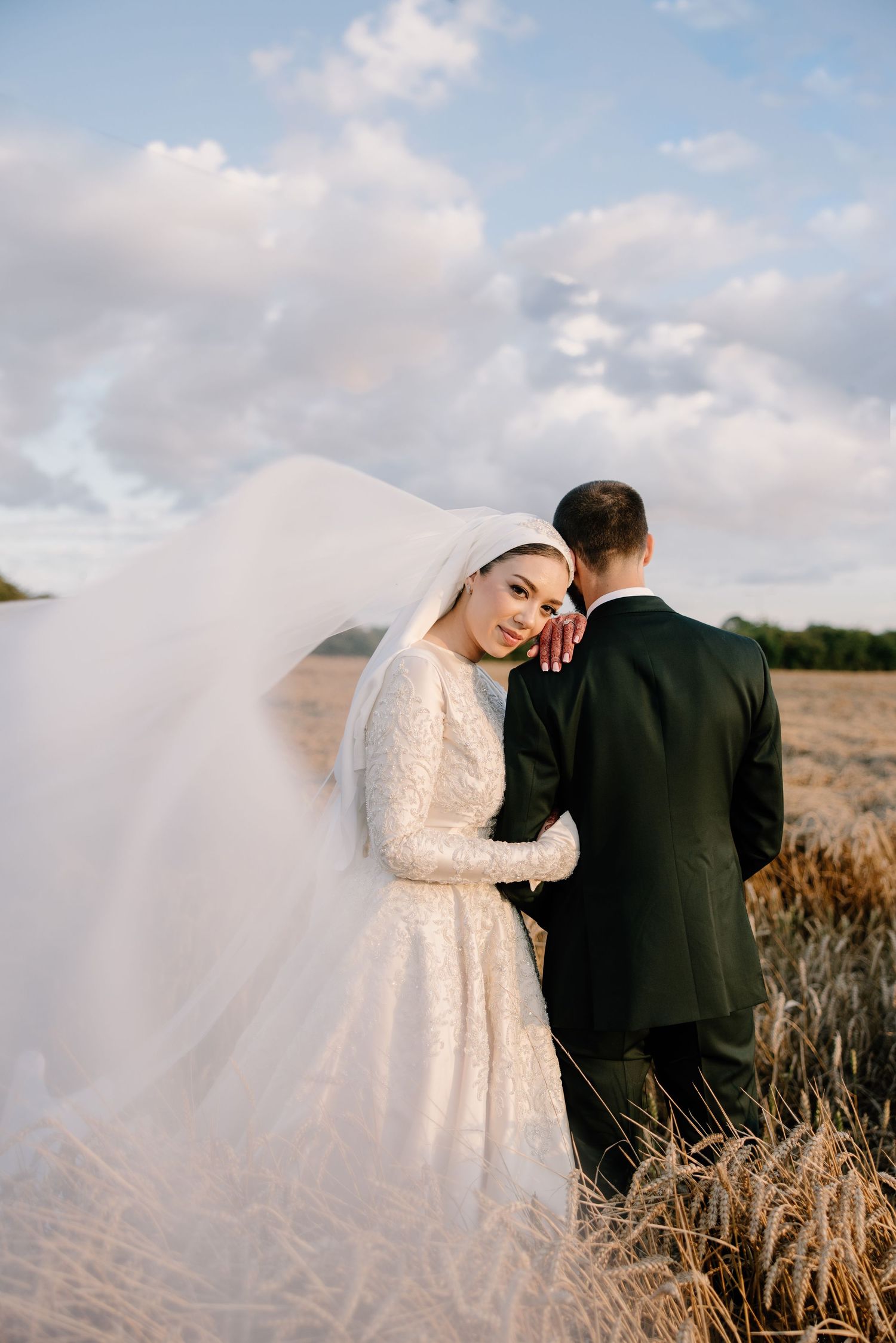 A romantic wedding portrait in a field with flowing veil and dramatic clouds at sunset.