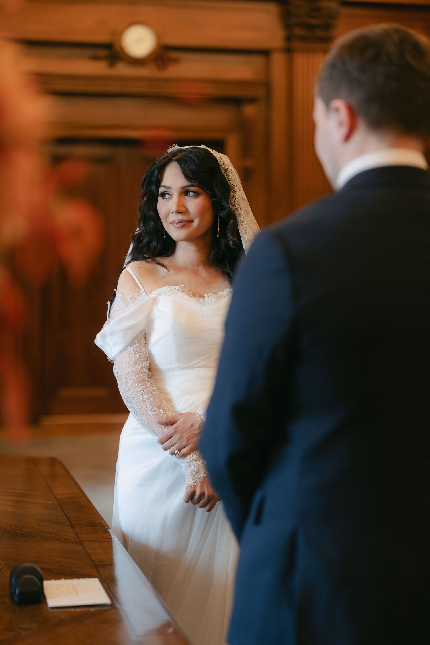 A bride in an elegant off-shoulder white wedding dress stands in a warmly lit indoor venue.