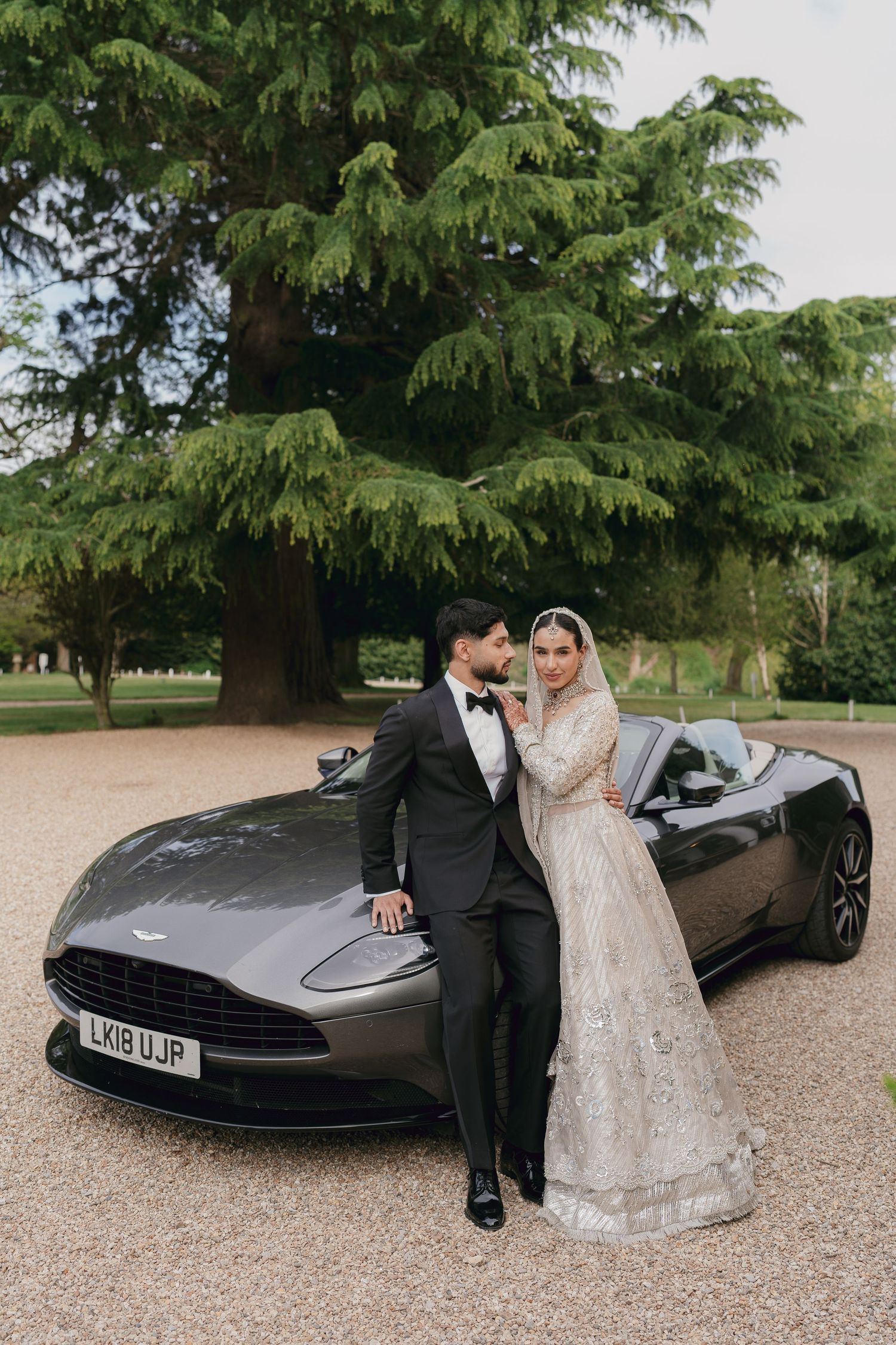 Bride and groom posing elegantly beside a grey Aston Martin DB11 on a gravel driveway surrounded by lush trees.