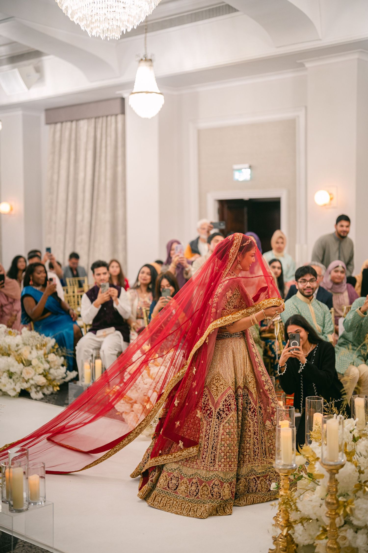 Traditional Indian wedding lehenga in red and gold displayed in an elegant white reception hall with chandeliers.