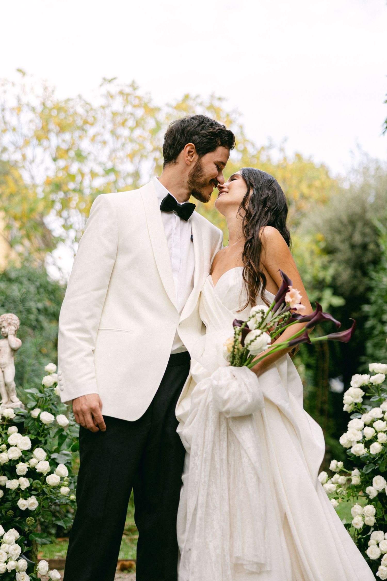 A romantic couple in wedding attire share intimate moments in a garden setting with white roses.