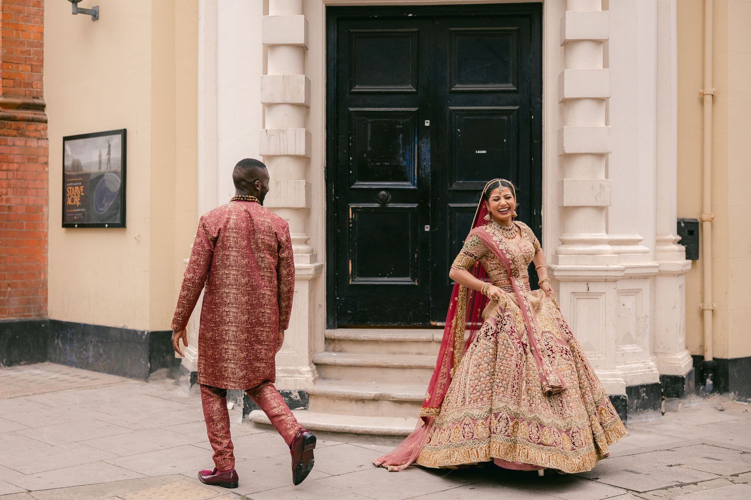 A couple in traditional Indian wedding attire walks down a street with cream-colored buildings and black doors.