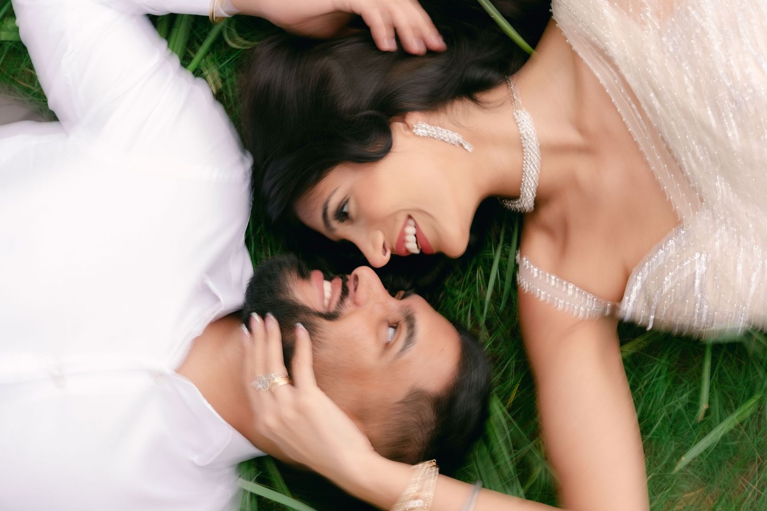 A joyful couple in wedding attire lying on green grass, smiling at each other in a romantic outdoor setting.