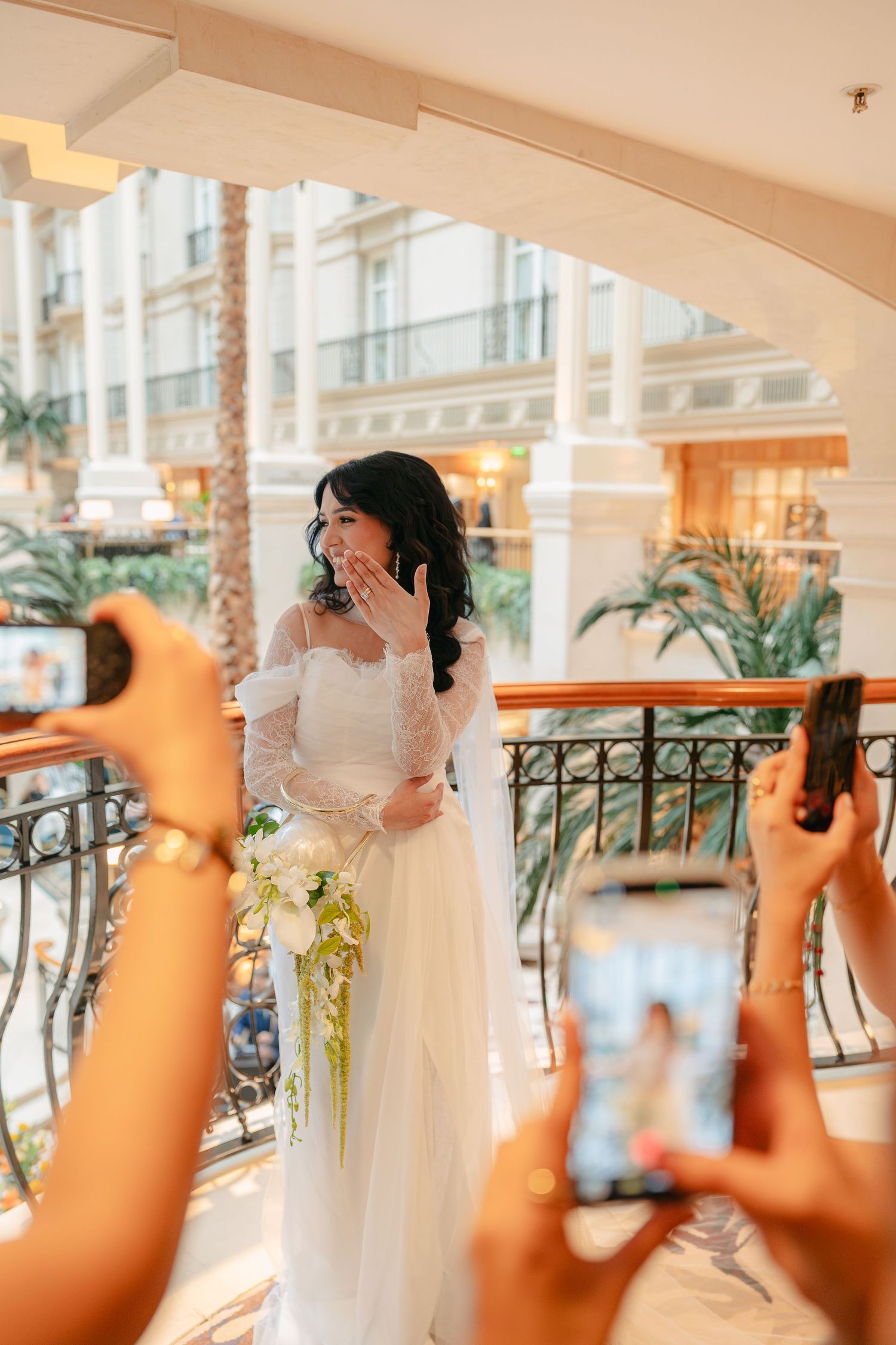 Bride in white lace gown holding bouquet poses on balcony while guests photograph her with smartphones.