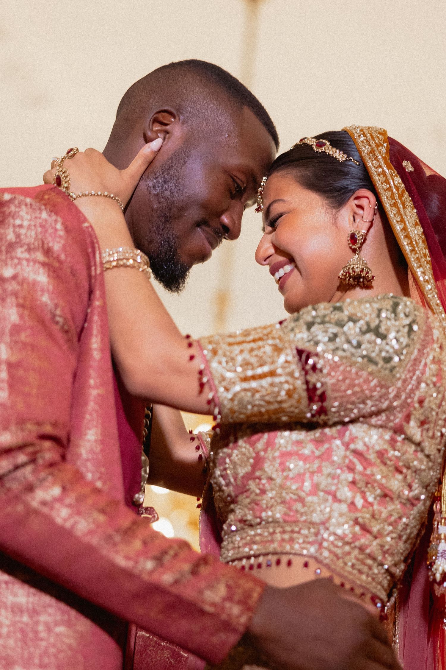 An intimate moment between newlyweds wearing traditional South Asian wedding attire in shades of red and gold.