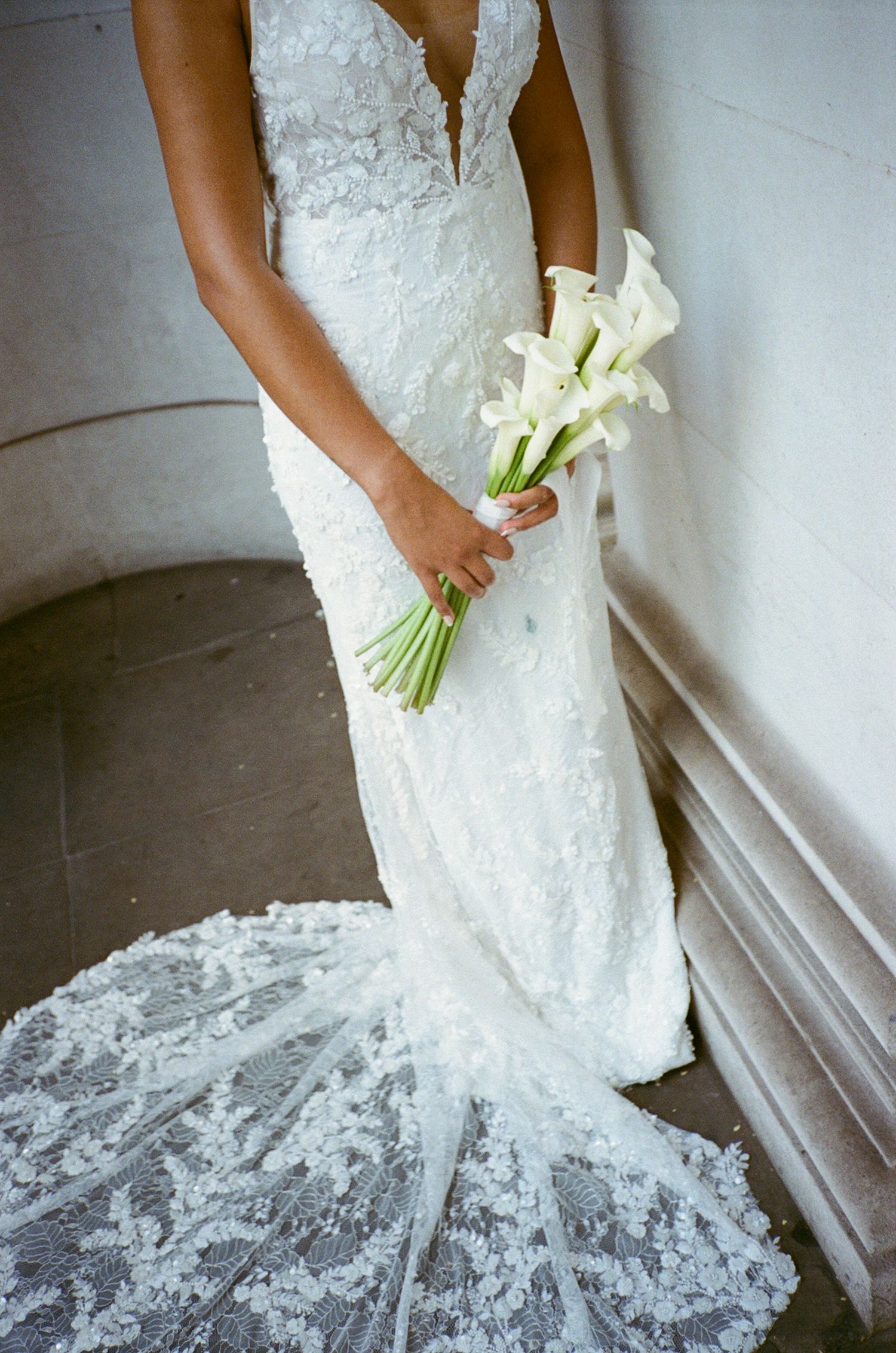 Bride in white lace wedding gown with train holds elegant calla lily bouquet on stone staircase.