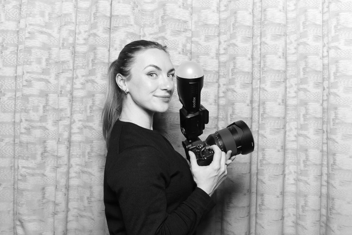 A photographer in black clothing holds a vintage camera while posing against a textured wall background.