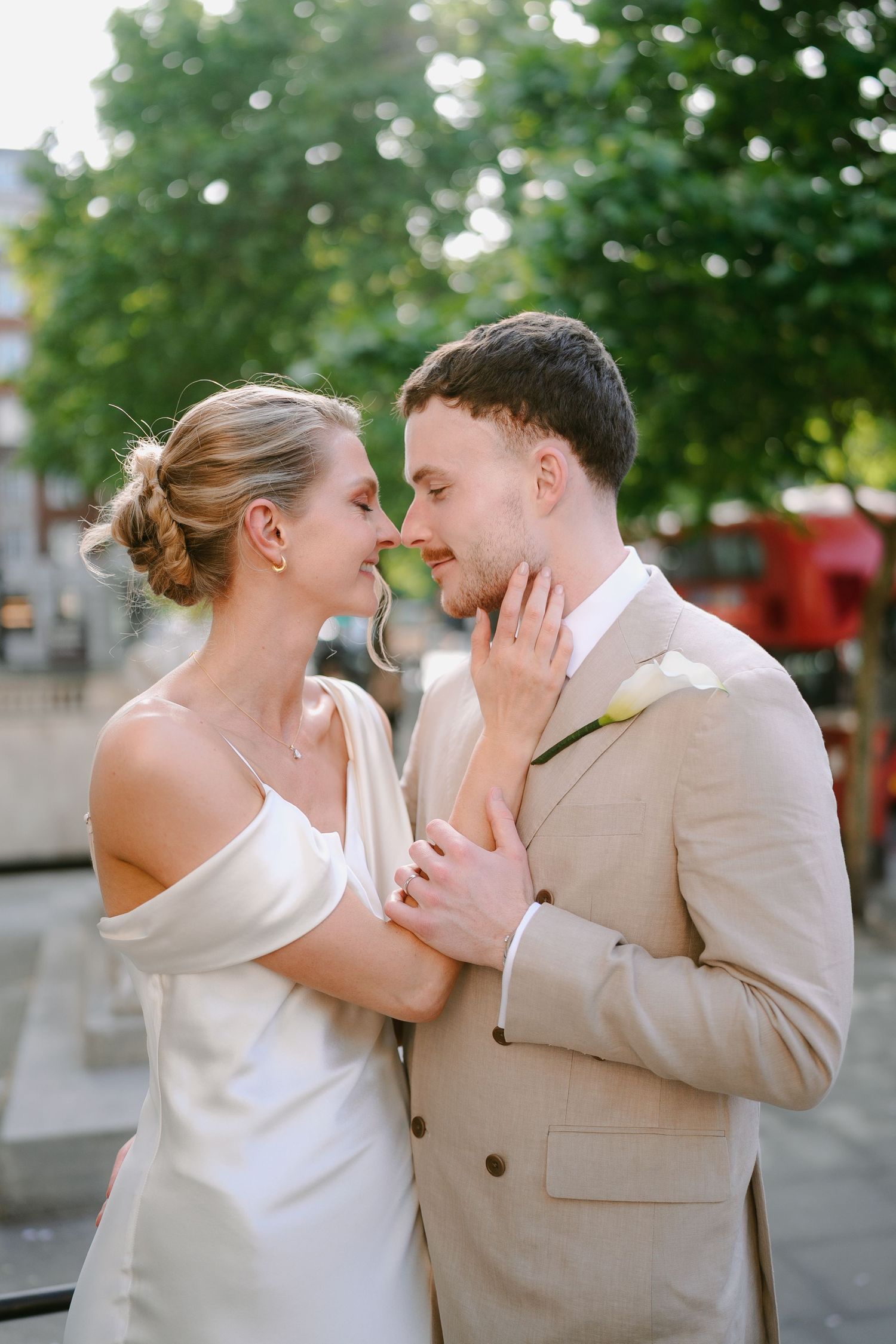A bride and groom share a romantic moment, nearly kissing outdoors, she in a white gown, he in a tan suit with a boutonniere.