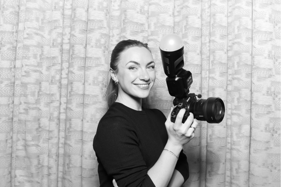 Professional photographer in black attire smiles while holding camera equipment against a textured wall background.