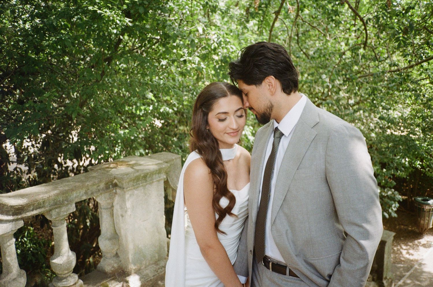 A couple shares an intimate moment outdoors, the groom in a gray suit leaning close to his bride in a white dress.