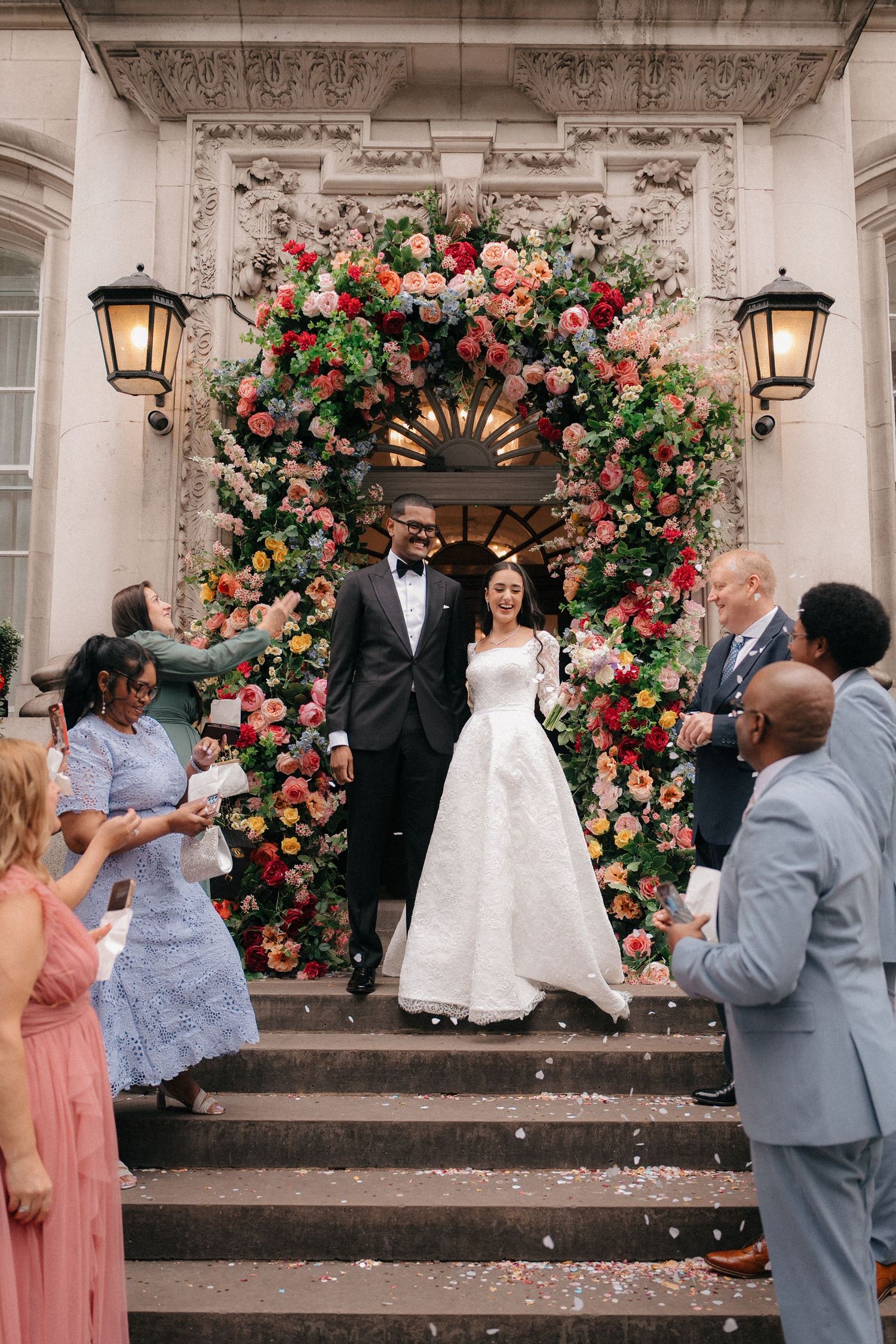 Wedding celebration outside historic building with beautiful floral arch and celebrating guests.