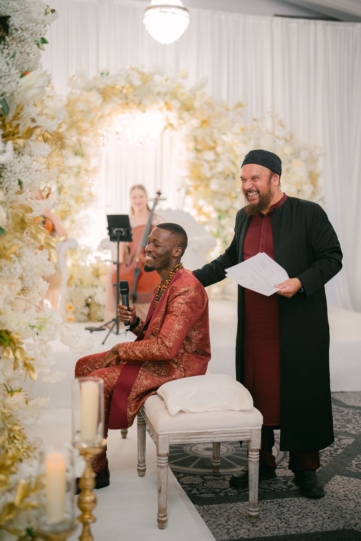 Wedding ceremony under a floral arch with traditional attire and decor in an elegant white venue.