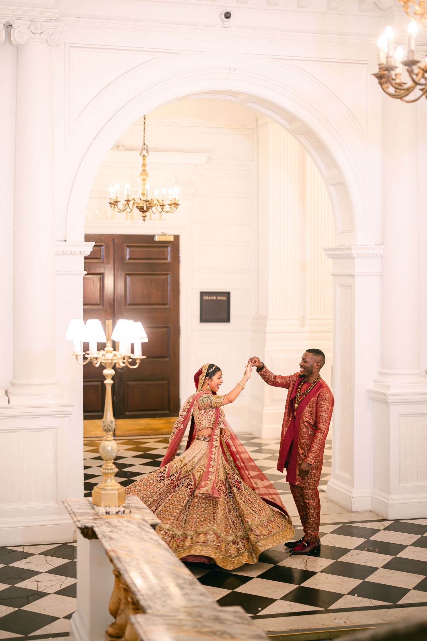 A couple in traditional Indian wedding attire dance together on a checkered floor beneath elegant chandeliers.