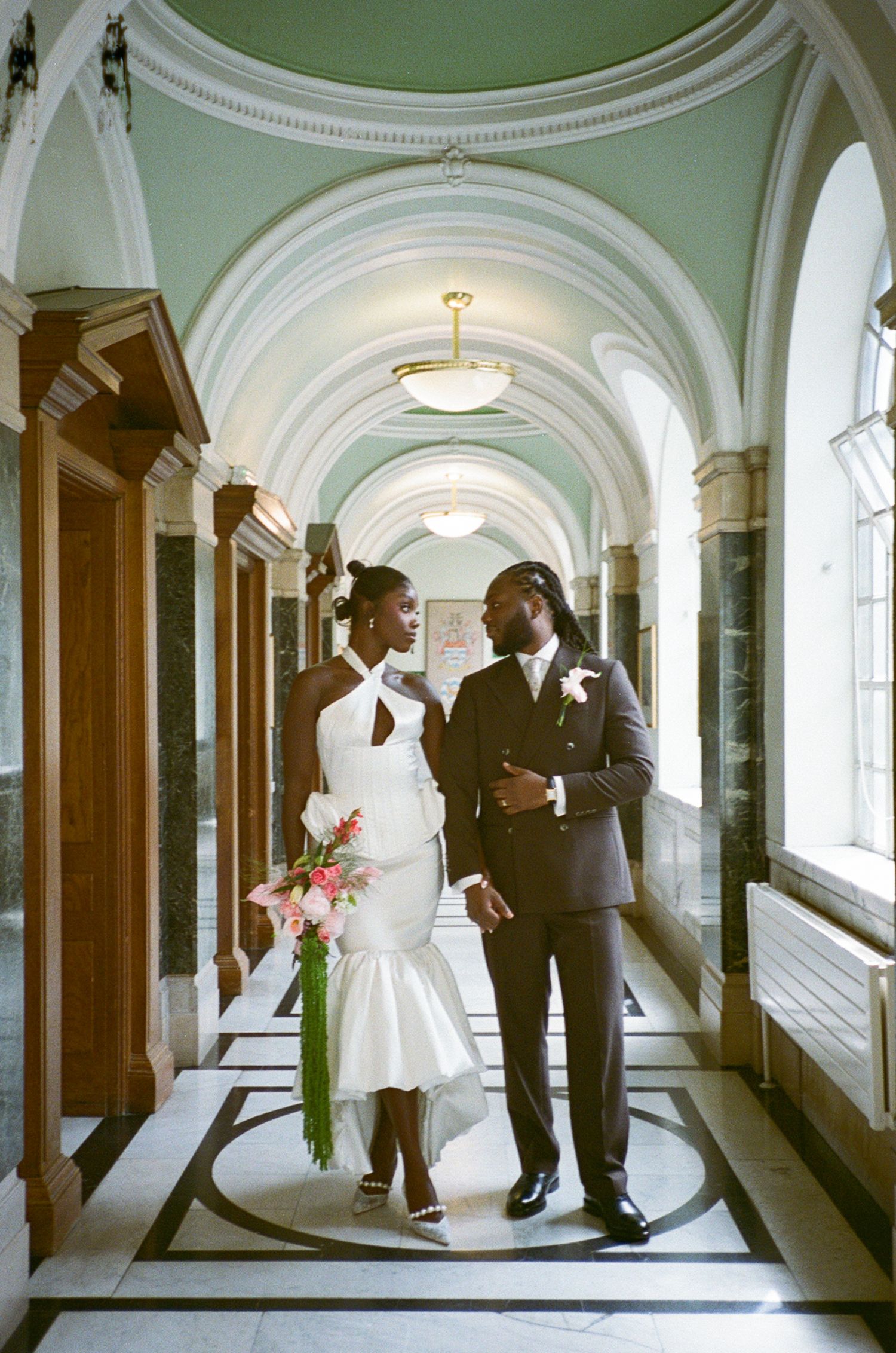 Elegant Black couple in wedding attire pose in ornate hall, bride in white gown holding flowers, groom in dark suit.