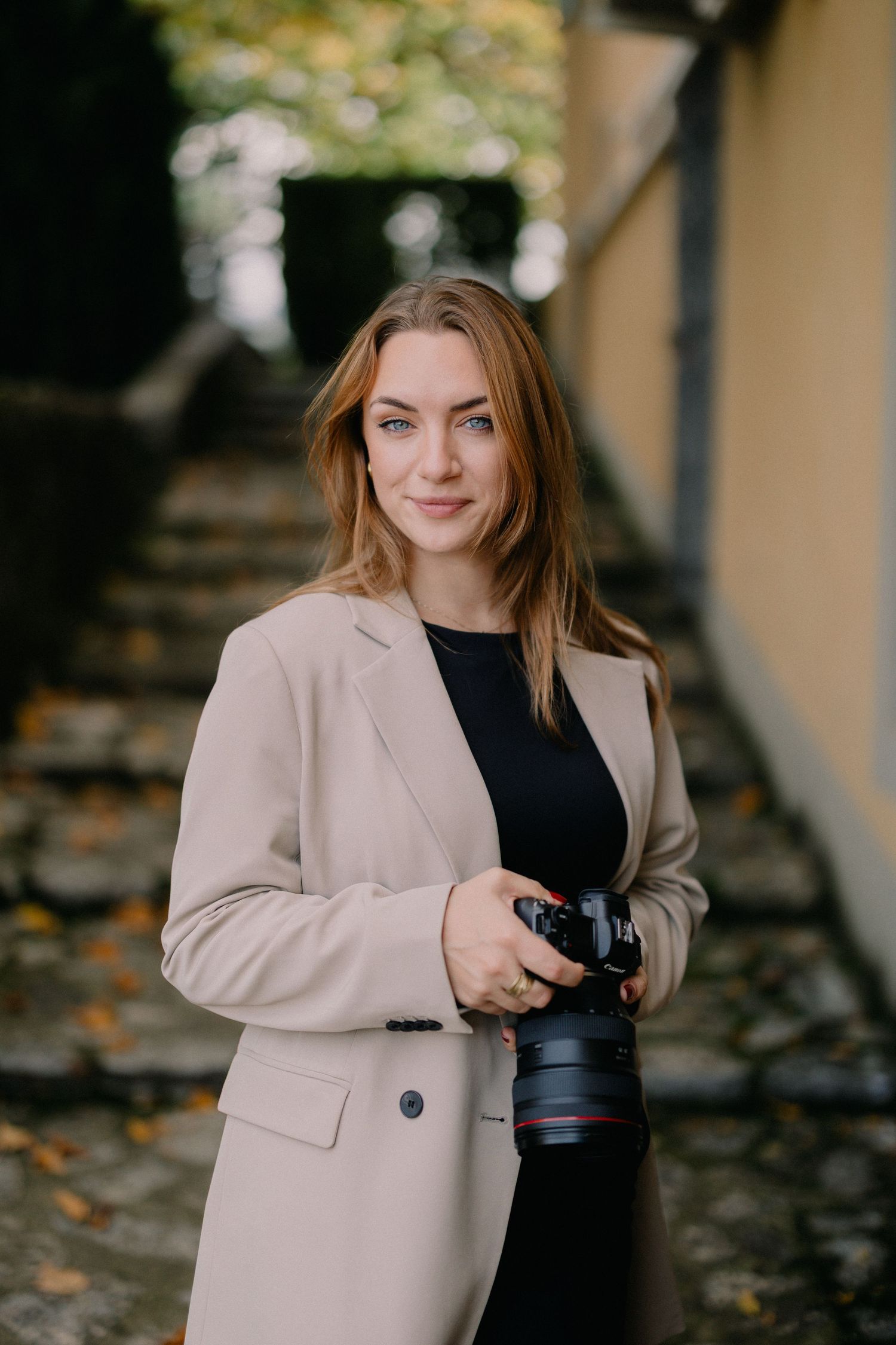 Professional photographer in beige blazer and black top holding camera on outdoor steps with autumn leaves.