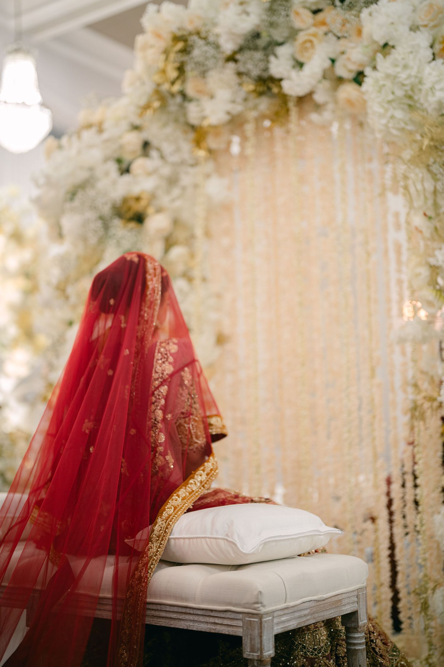 Traditional Indian wedding decor with red veil and floral arrangements on white seating against elegant backdrop.