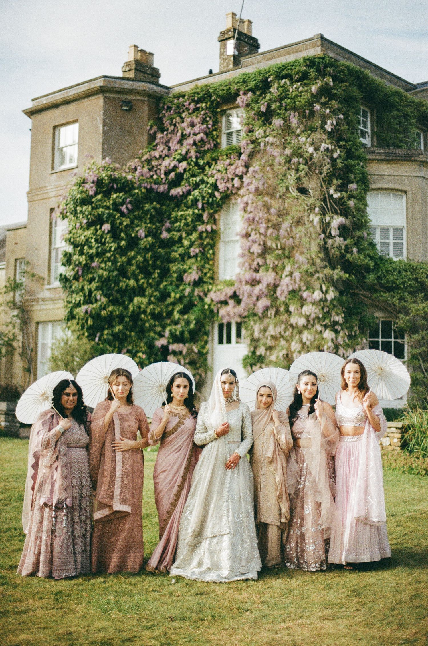 Bride in white lehenga with bridesmaids in pink sarees holding parasols outside ivy-covered English manor house.