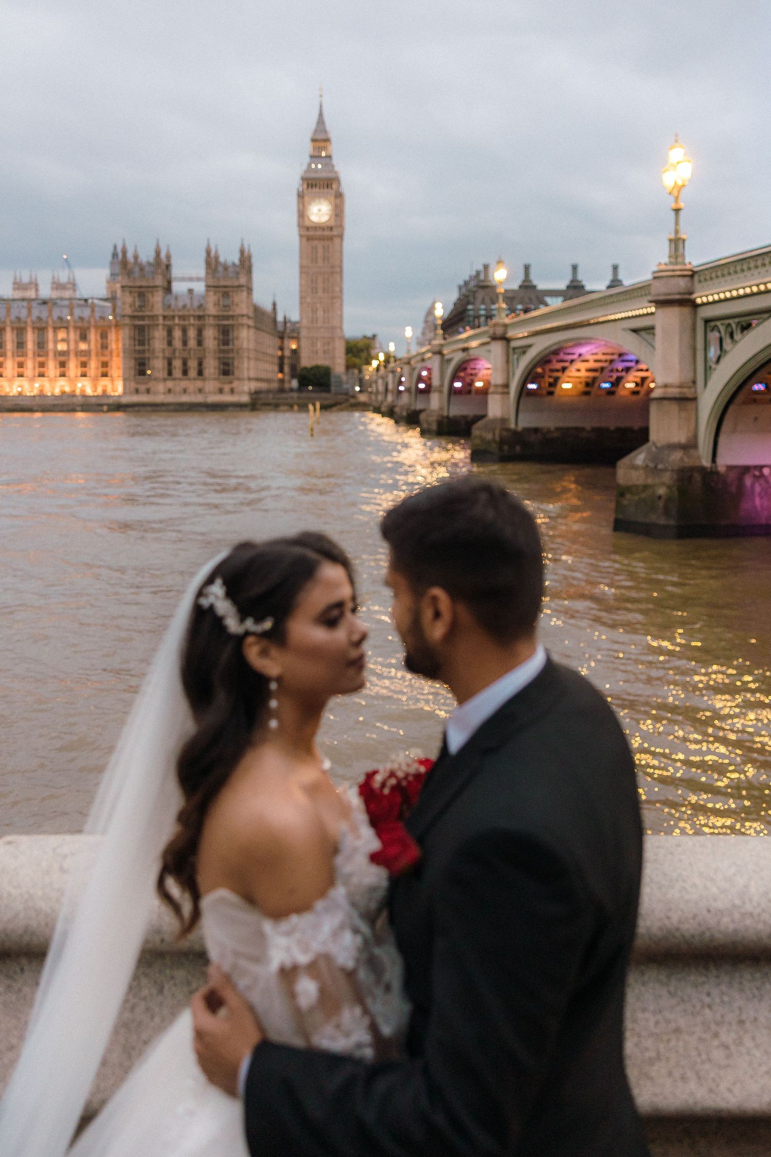 Bride and groom share a romantic moment on the Thames with Big Ben and Westminster Bridge illuminated at dusk.