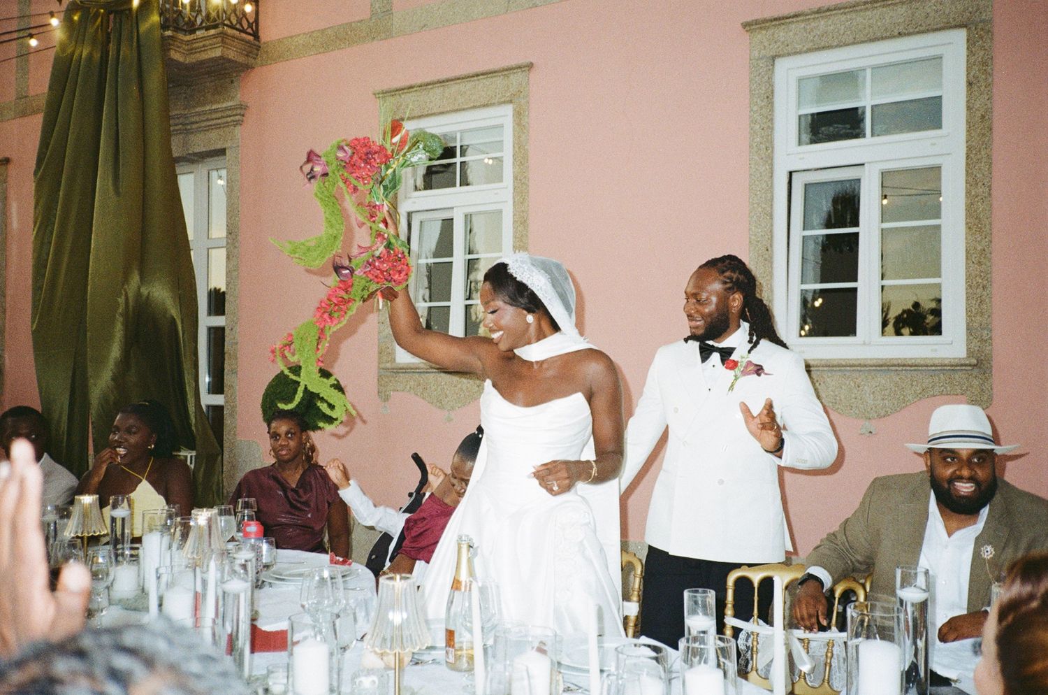 Black bride in white dress raises floral bouquet while groom in white suit smiles during wedding reception dinner.