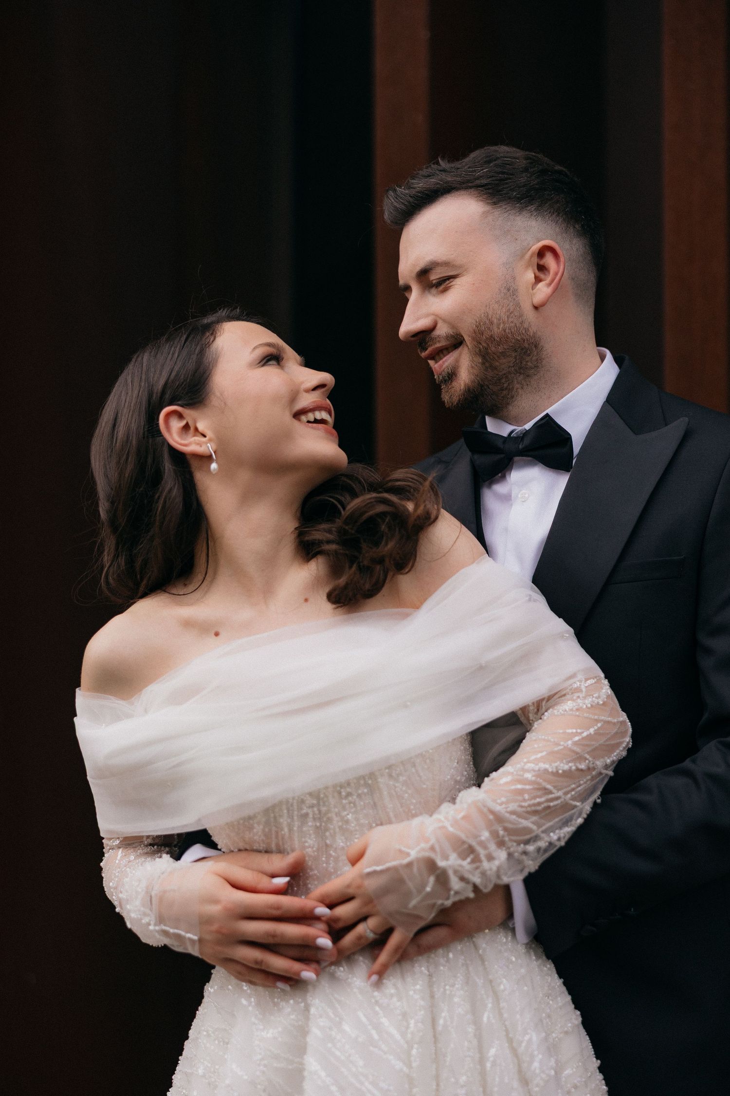 Bride in lace dress laughing while embraced by groom against dark background.