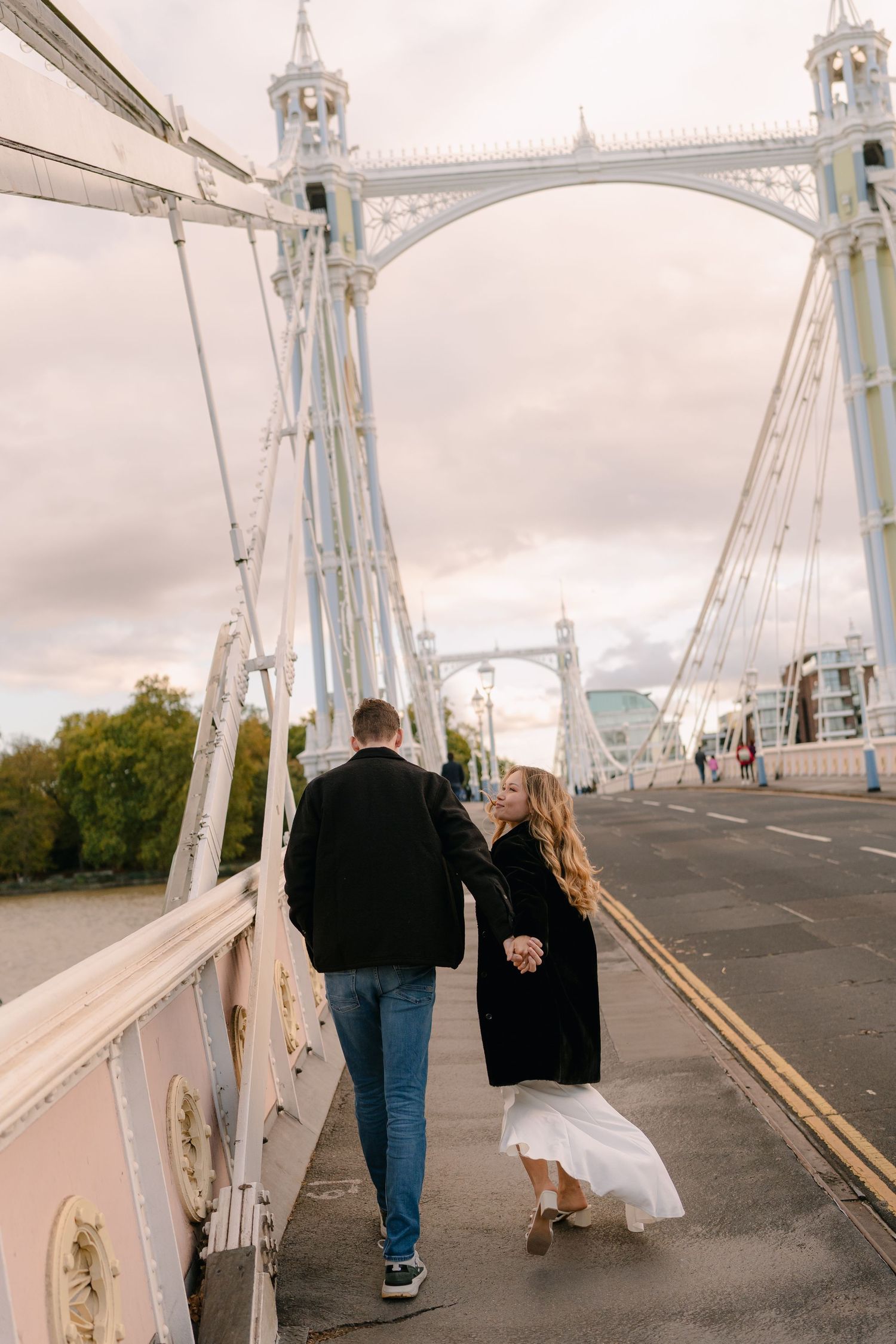 A couple walks along a white suspension bridge on an overcast day.