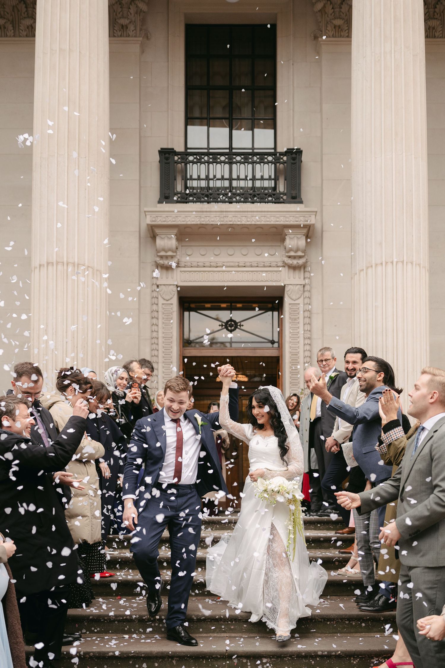 Bride and groom exit wedding venue as guests throw white confetti on grand stone steps with tall columns.