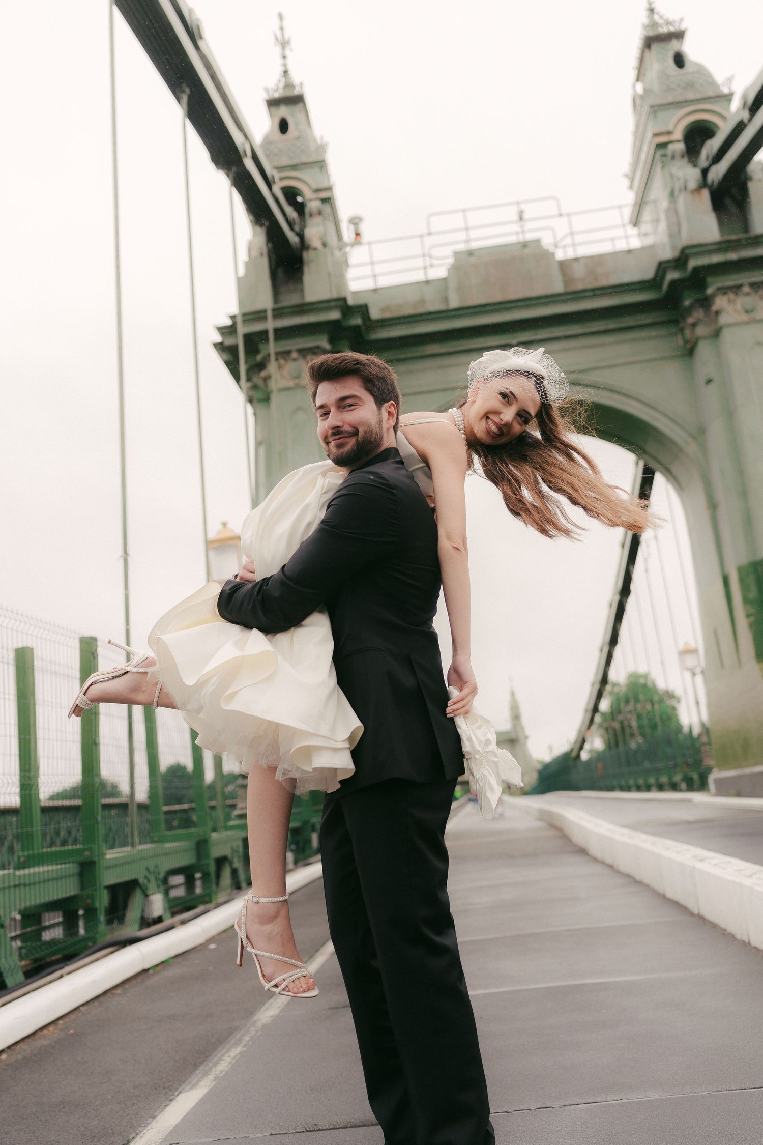 Groom carrying bride in white dress on a green bridge, laughing together on their wedding day.