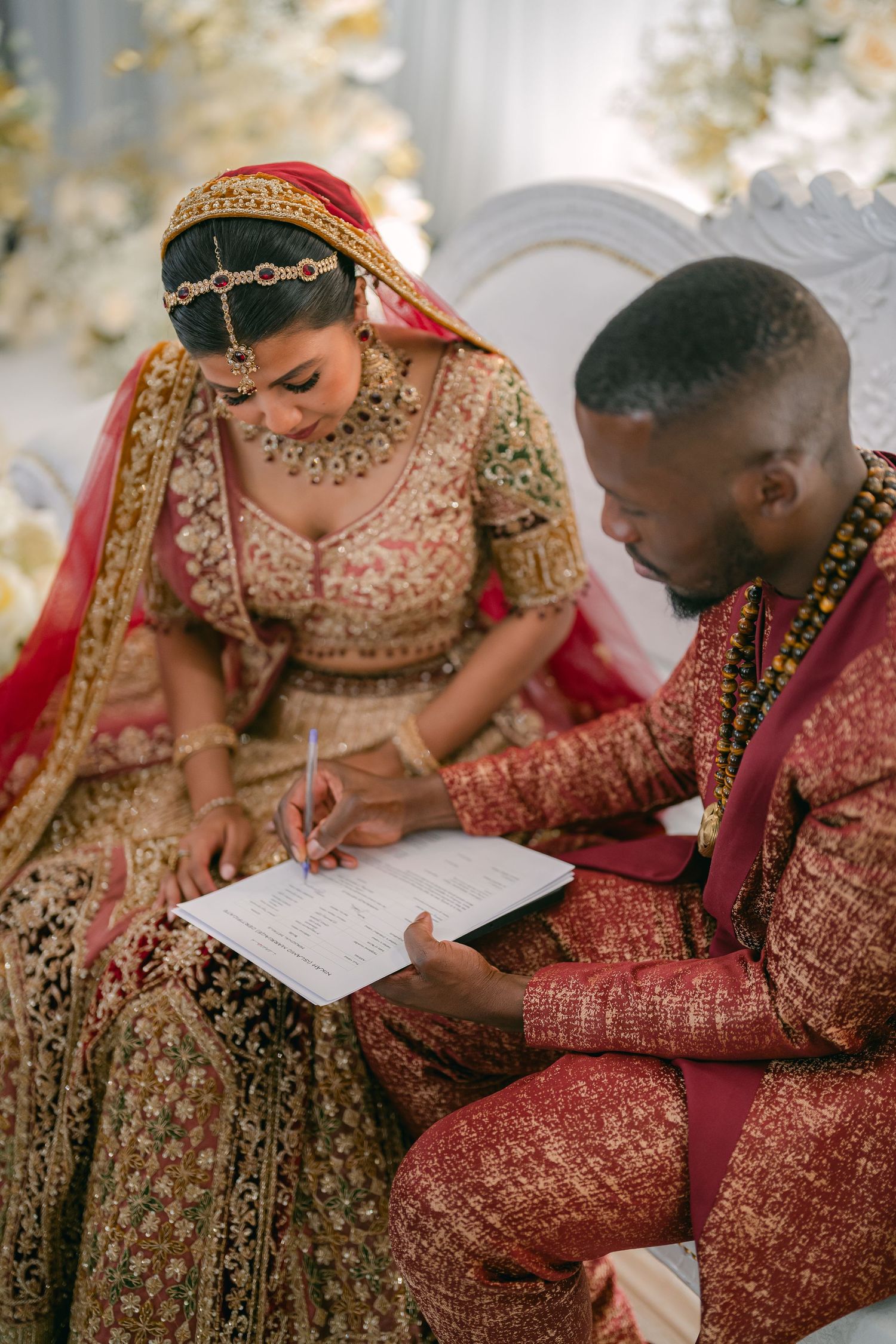 A couple in traditional Indian wedding attire signs marriage documents together in an elegant setting.