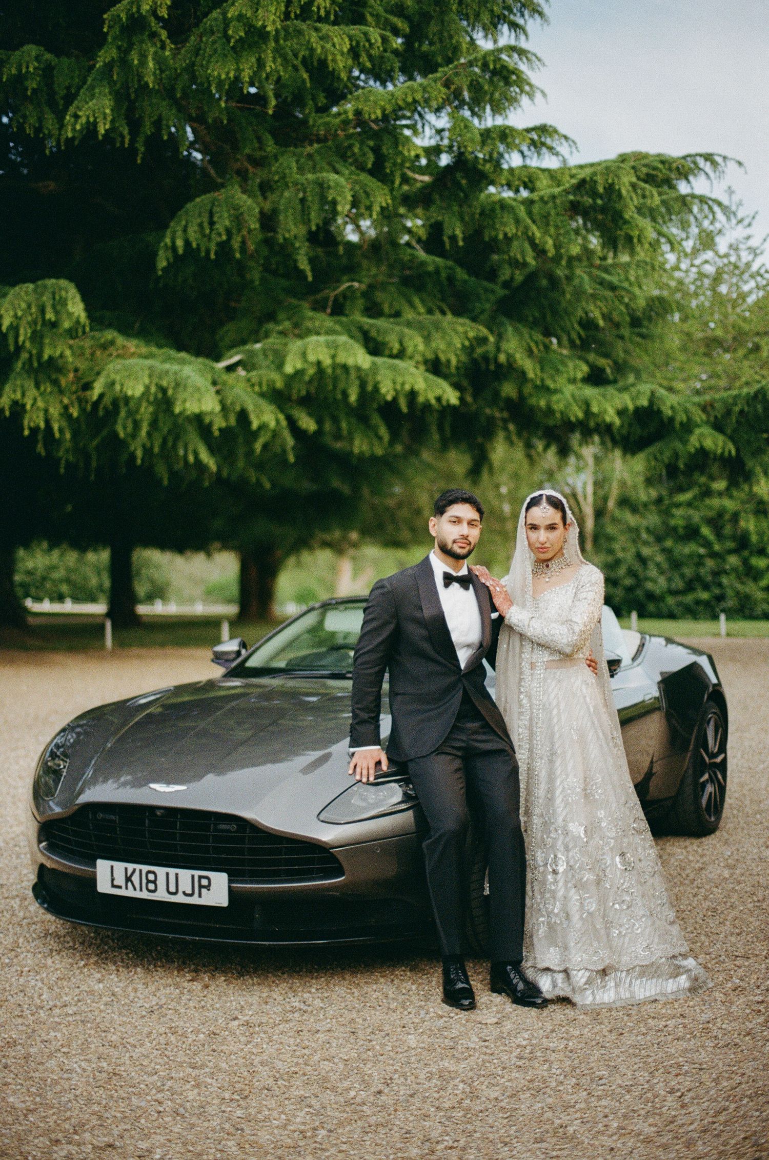Bride and groom posing beside a grey Aston Martin DB11 on a gravel driveway surrounded by lush trees.