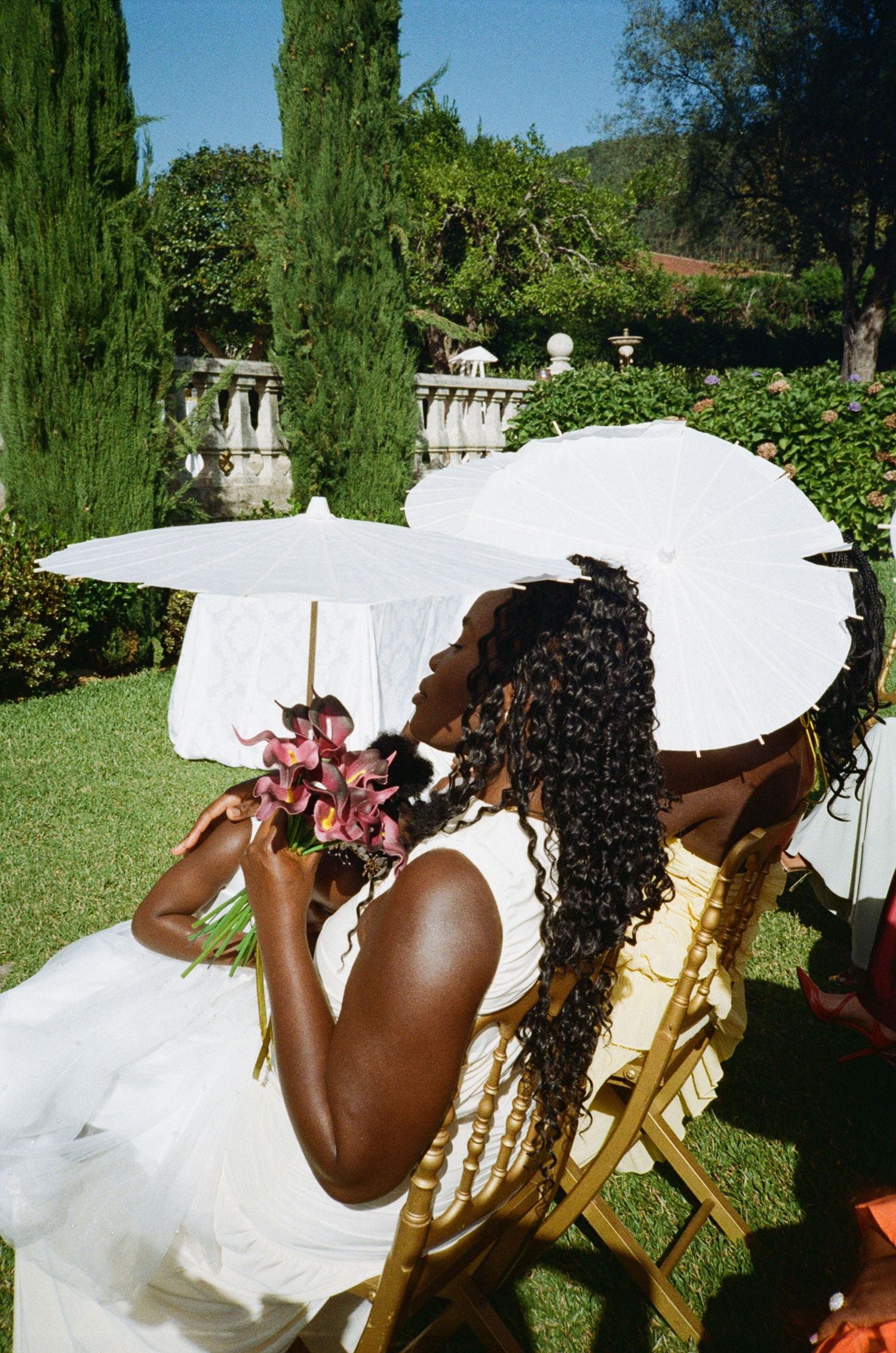 A Black bride in a white dress holds flowers under a white parasol while seated in a garden chair outdoors.