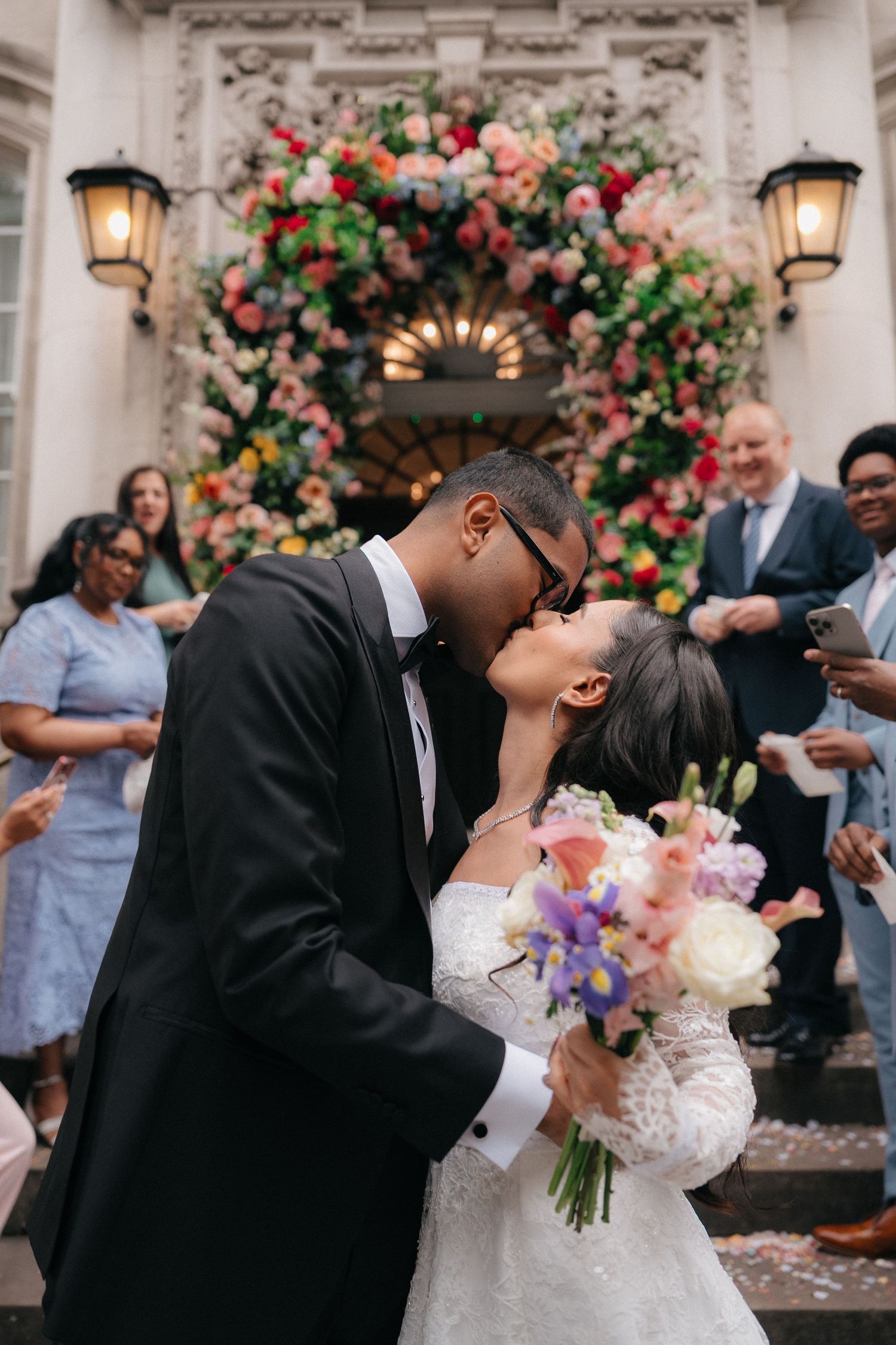 Romantic wedding kiss under a spectacular floral arch with guests celebrating.