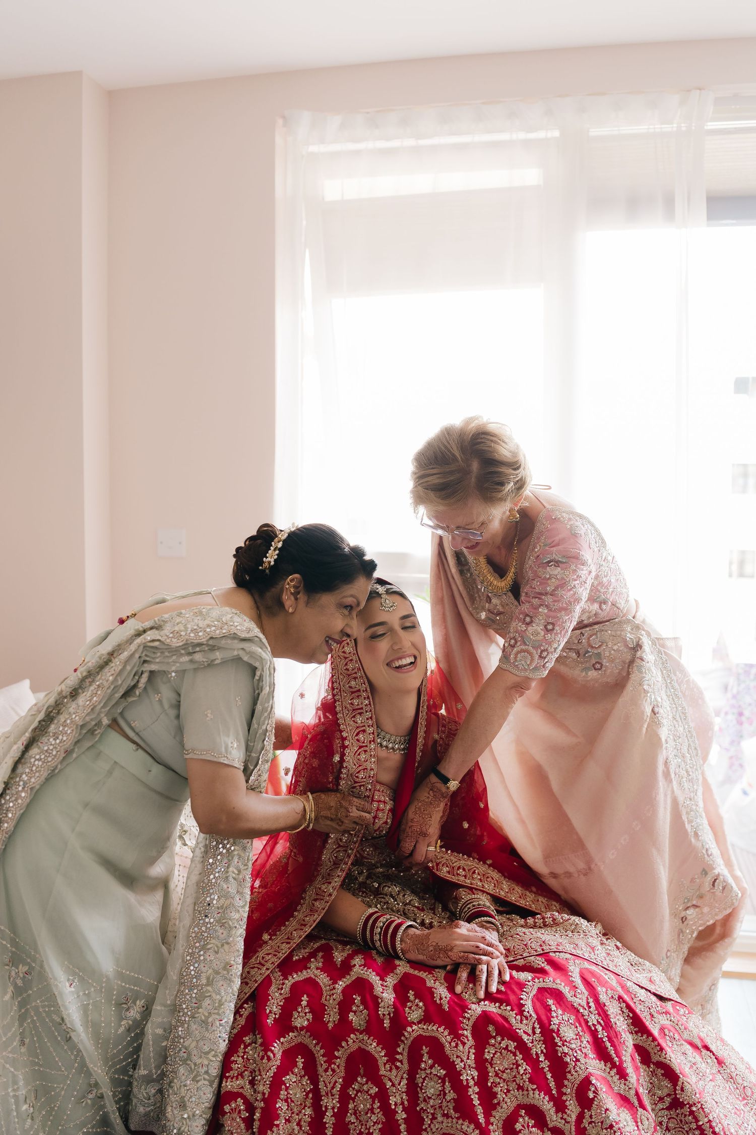 Bride in red lehenga laughing with two bridesmaids in silver and pink sarees during wedding preparations.
