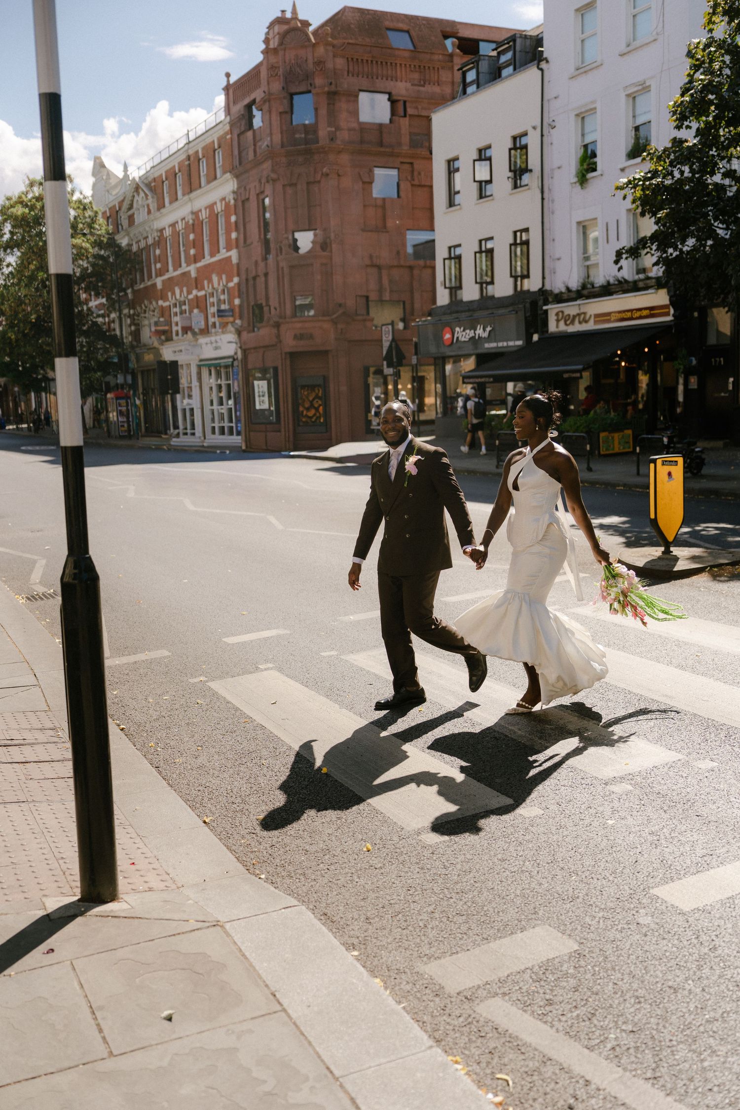 Newlywed couple holding hands crossing a zebra crossing on a sunny urban street, bride in white dress holding bouquet.
