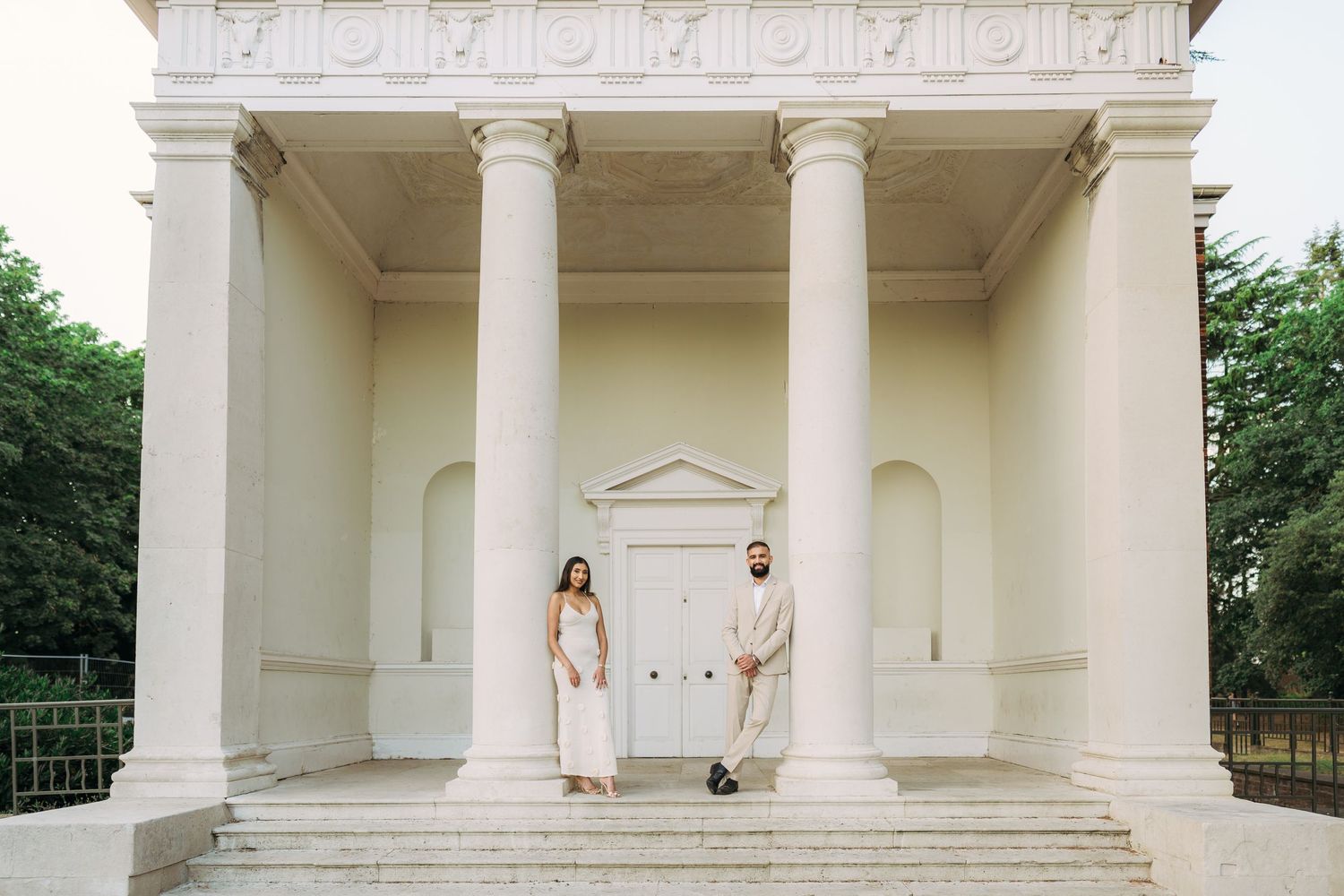 Couple standing on steps of white neoclassical building with tall columns, dressed in elegant attire for a wedding shoot.